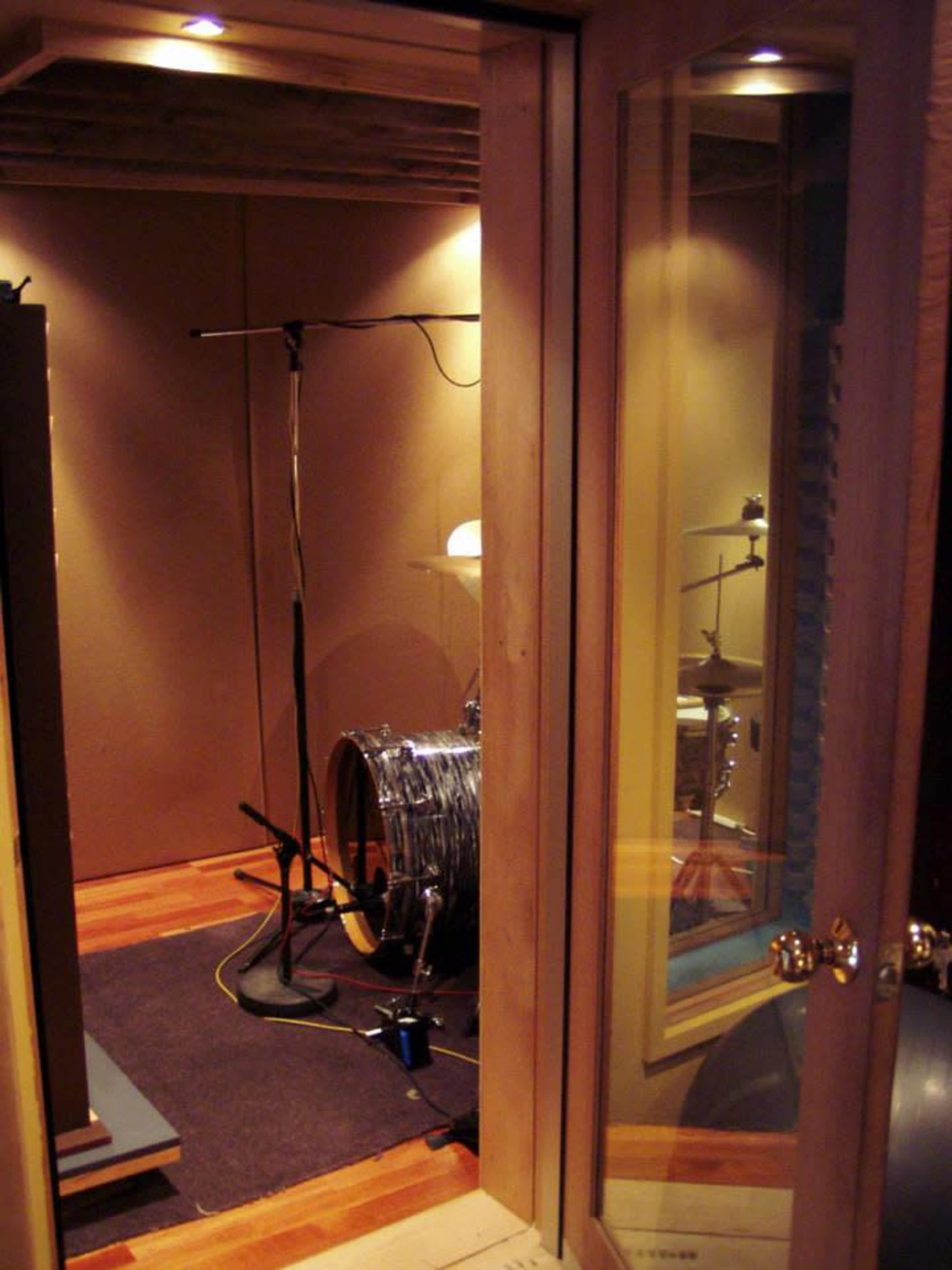 A small soundproof recording booth with a drum set partially visible through a glass door.