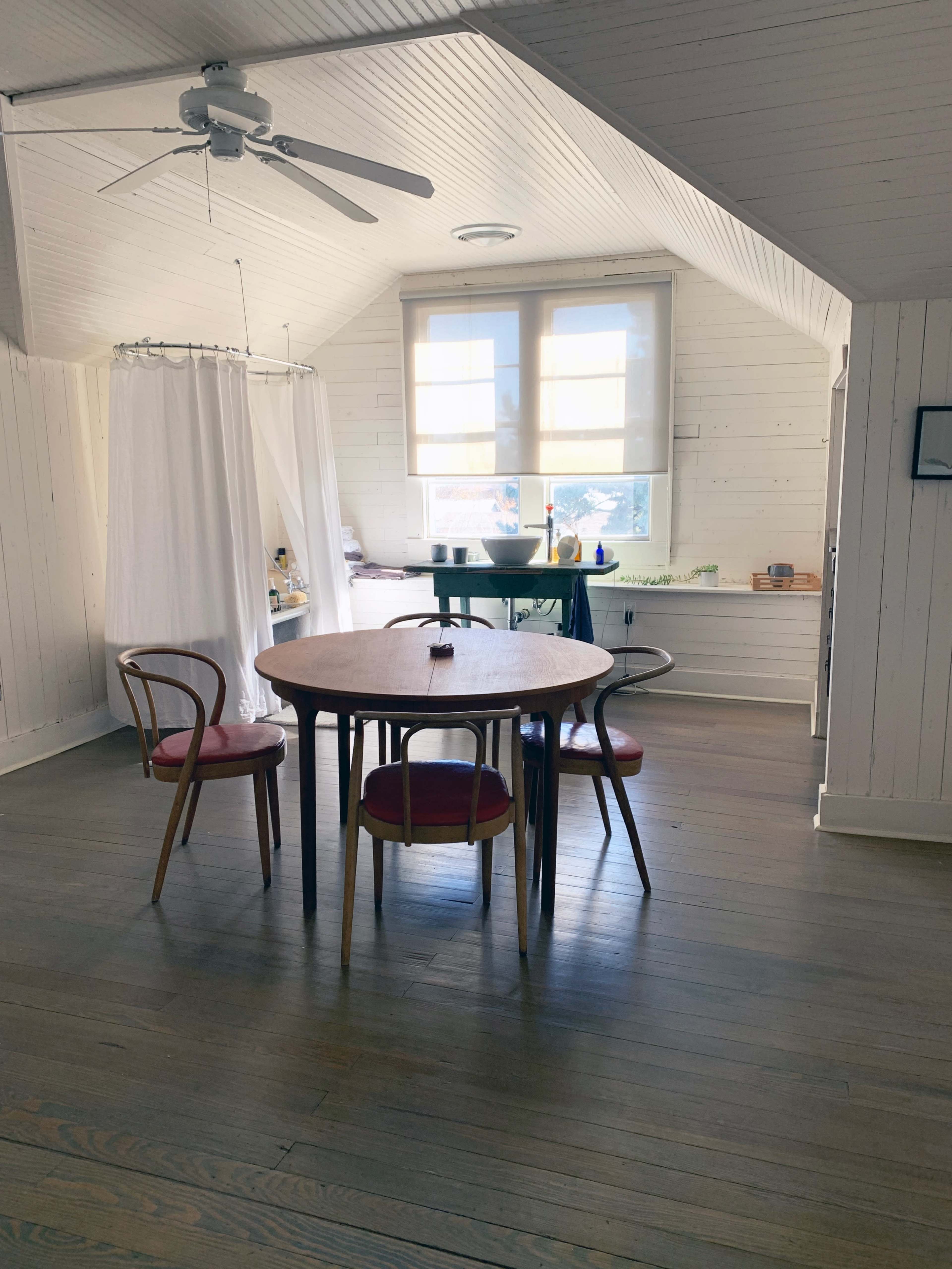 A wooden table with four chairs is situated in a room featuring white walls and large windows that allow natural light to enter.