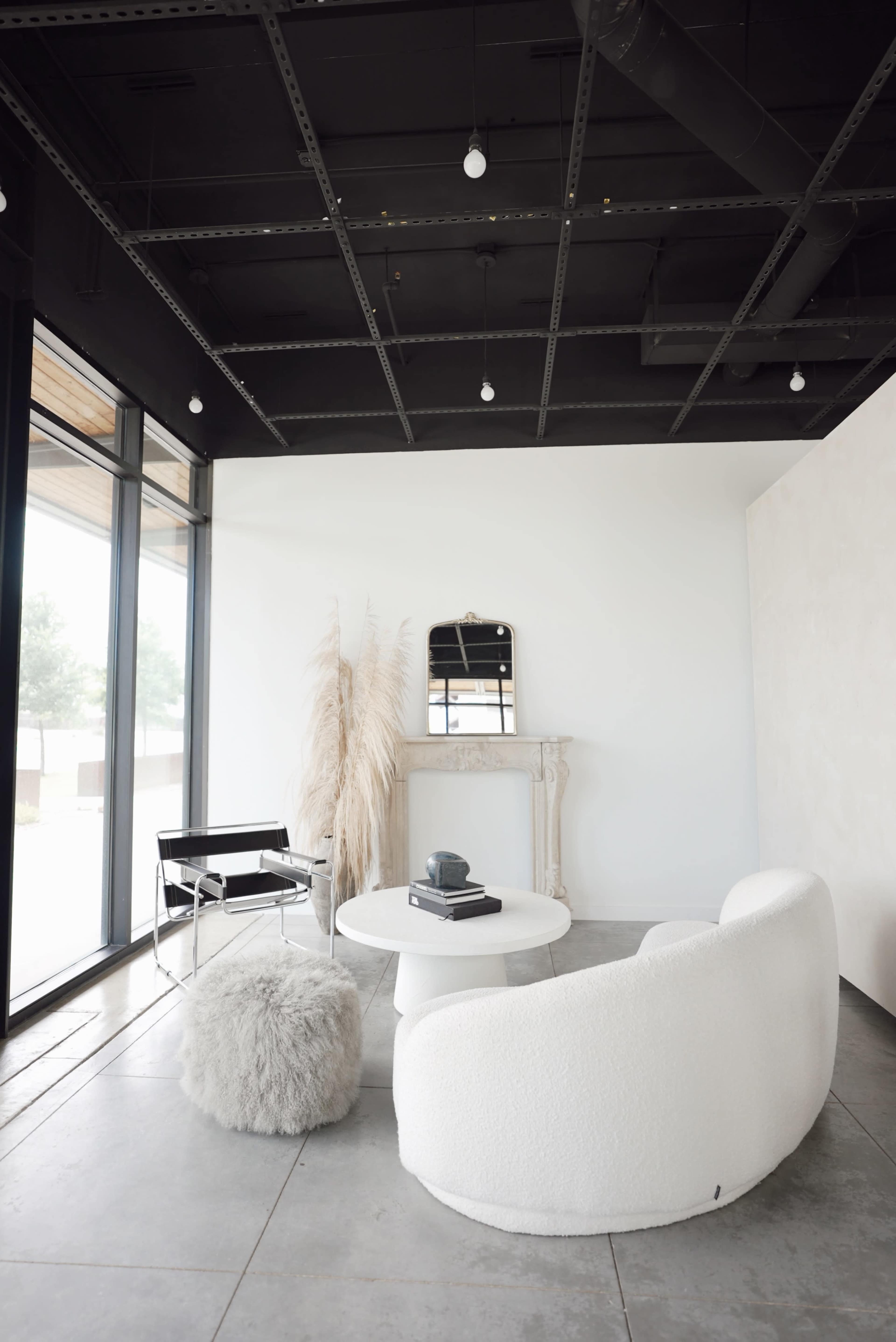 The scene features a minimalist interior with a round white table, a fluffy gray ottoman, a curved white sofa, and large windows letting in natural light.