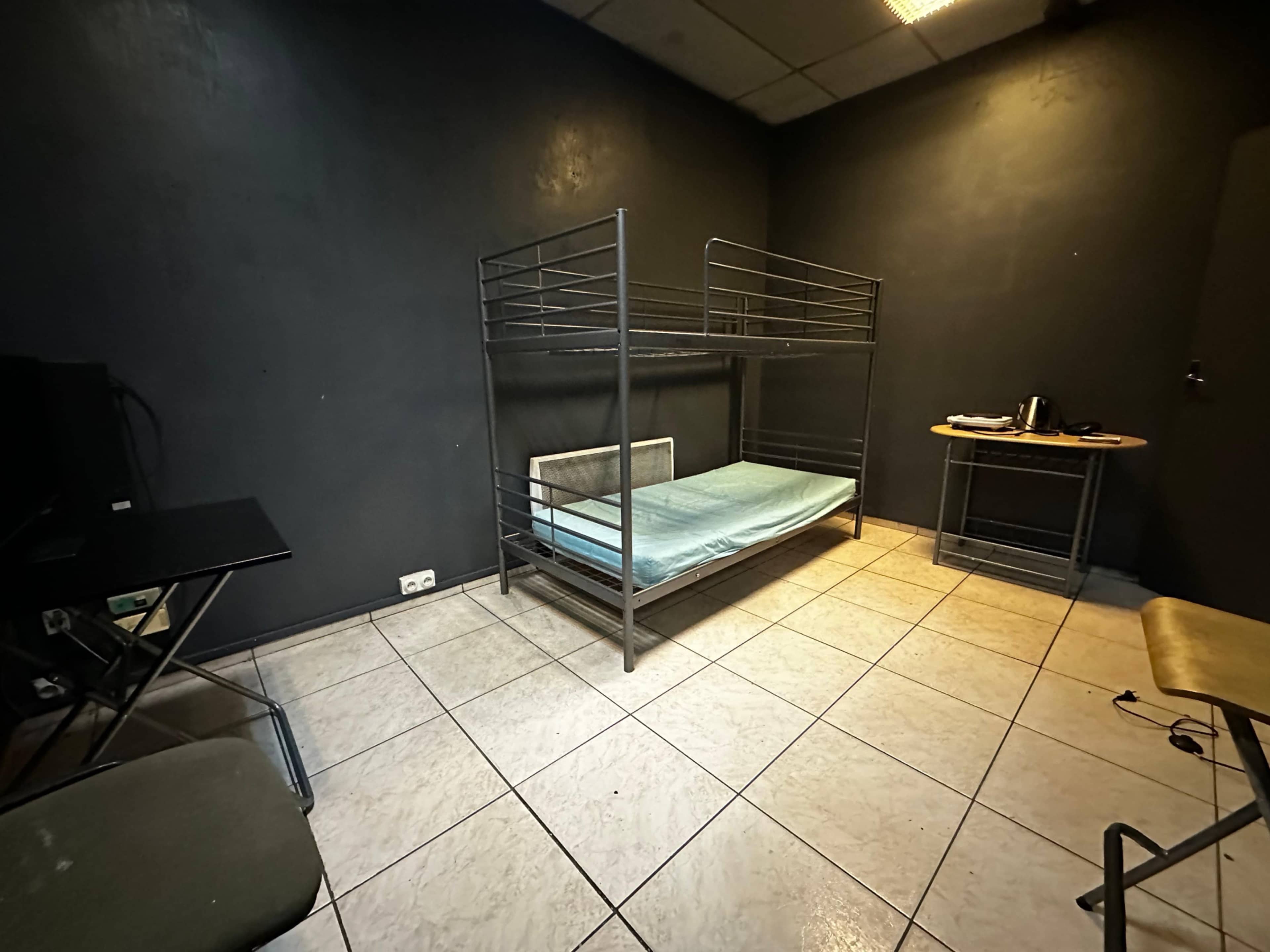The image shows a sparsely furnished room with a metal bunk bed on one side, a small table, and a chair in a corner.