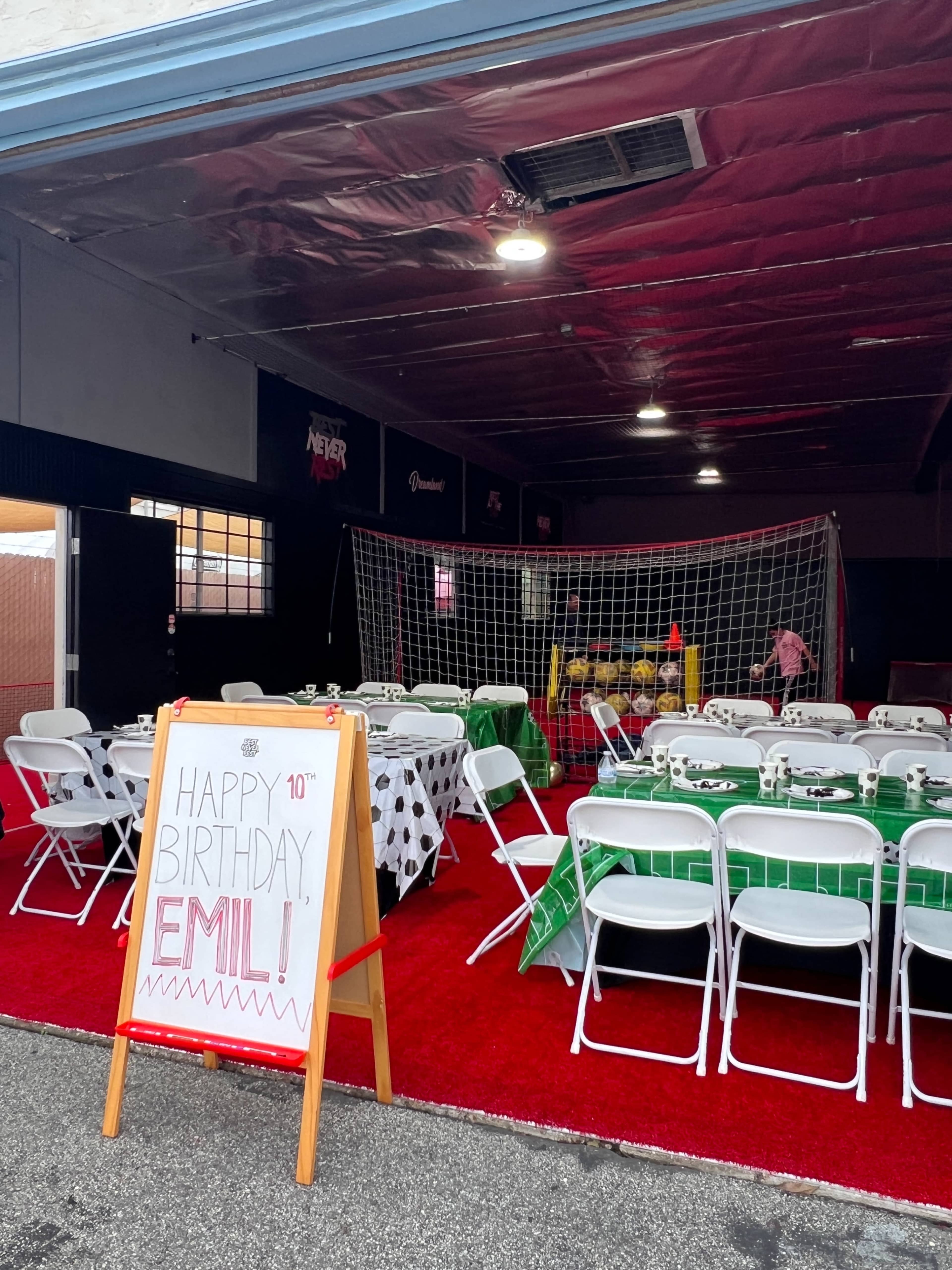 A birthday celebration setup is arranged in a garage, featuring decorated tables, chairs, and a soccer goal in the background.