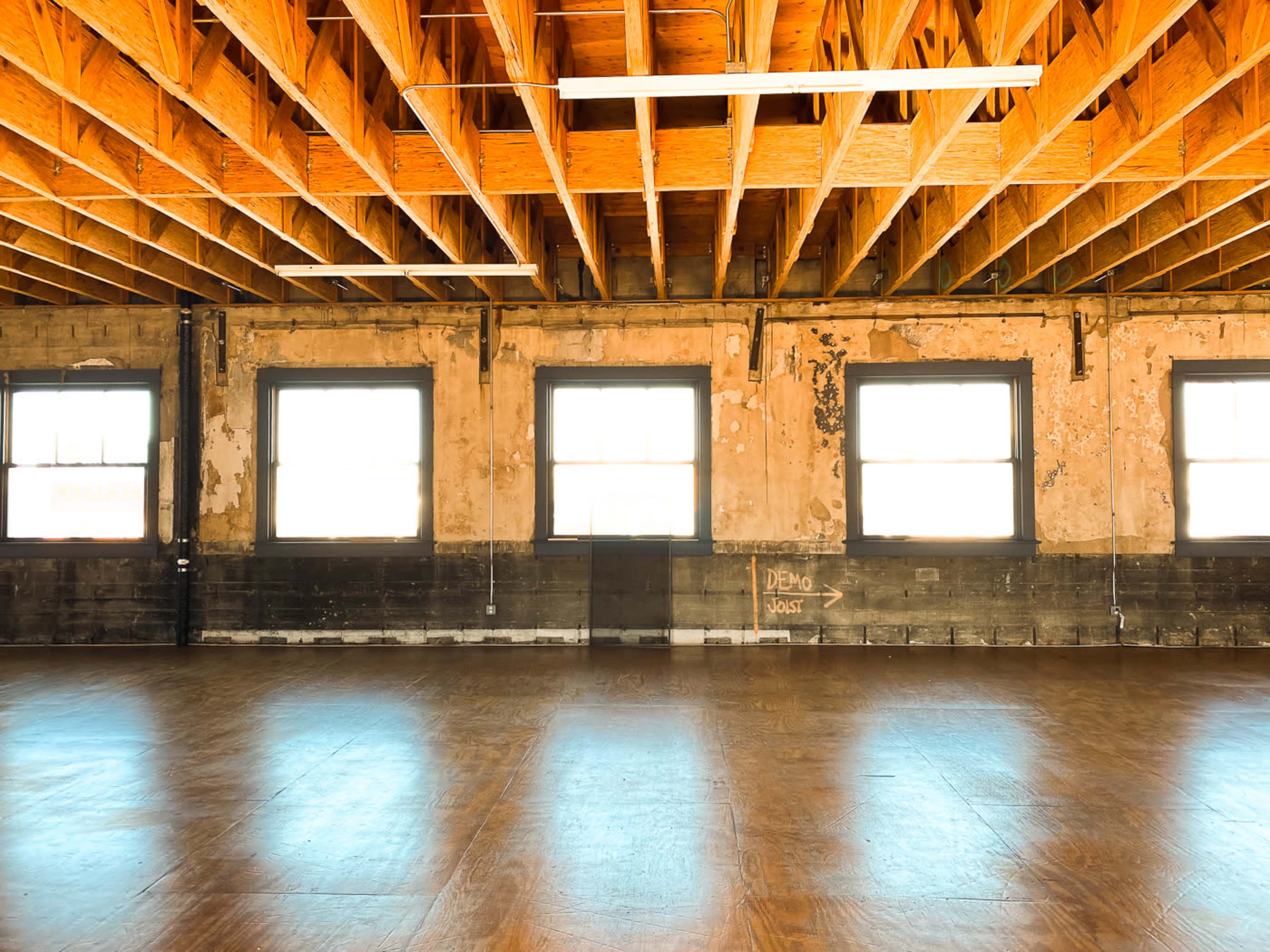 The image shows an empty room with wooden flooring, large windows, and exposed wooden beams in the ceiling.
