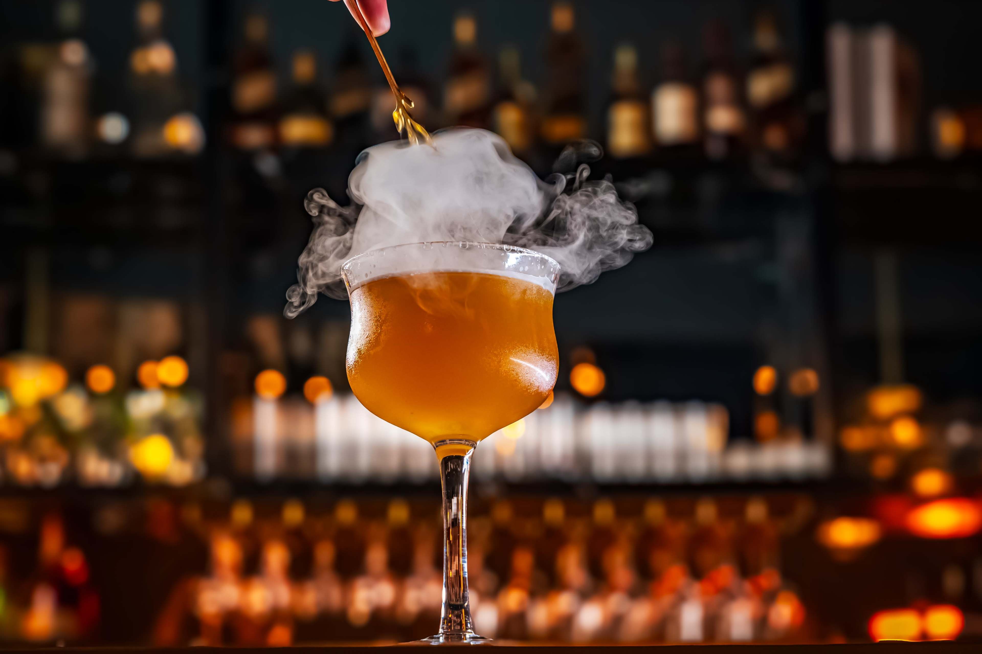 A cocktail glass with a smoky mist rising from it is prominently displayed against a blurred bar backdrop filled with bottles.