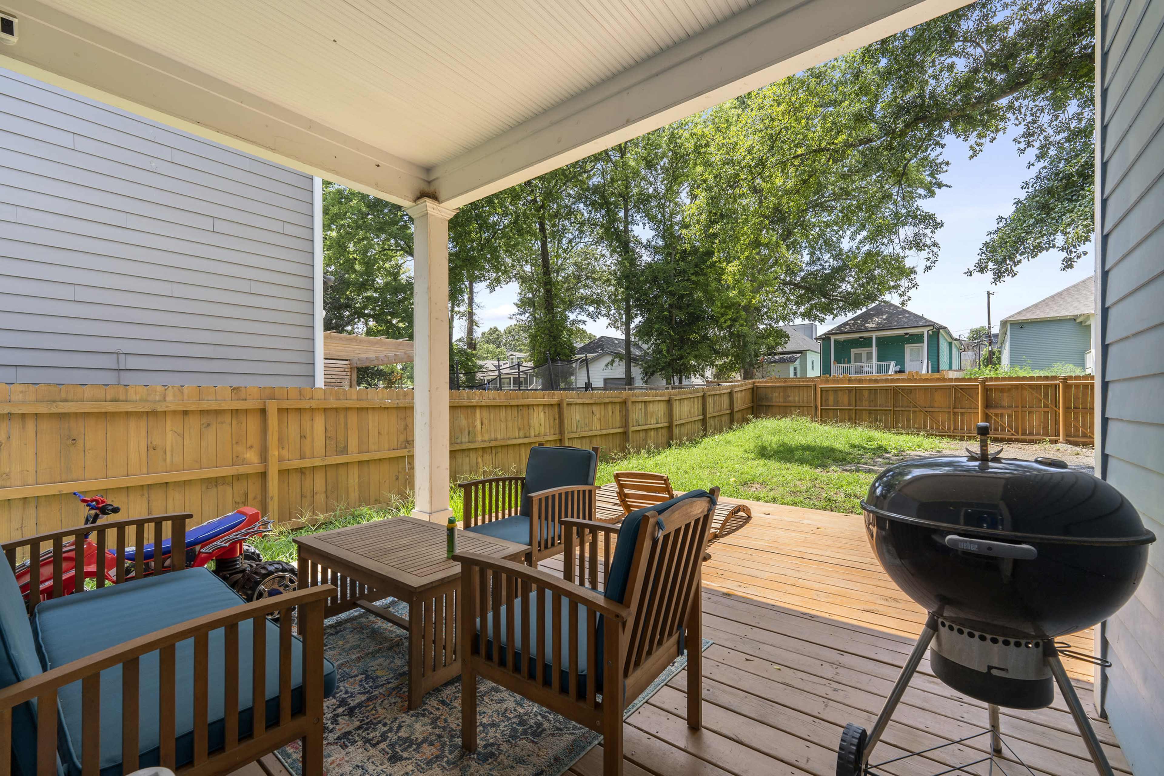 A covered patio features a wooden table and chairs, with a charcoal grill and a fenced backyard visible in the background.