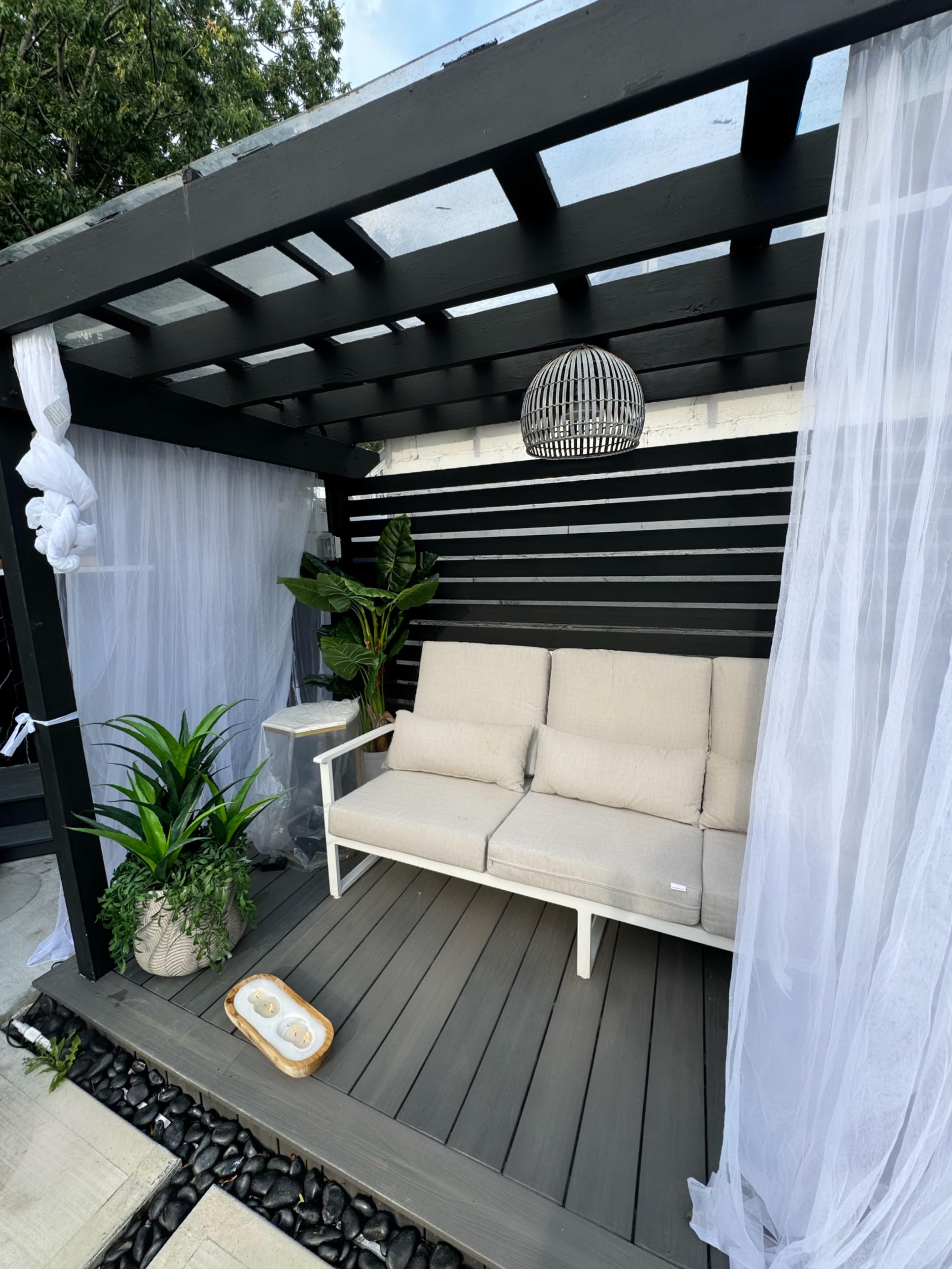 The image shows a modern outdoor seating area with a black wooden structure, two beige armchairs, a side table, potted plants, and sheer white curtains.