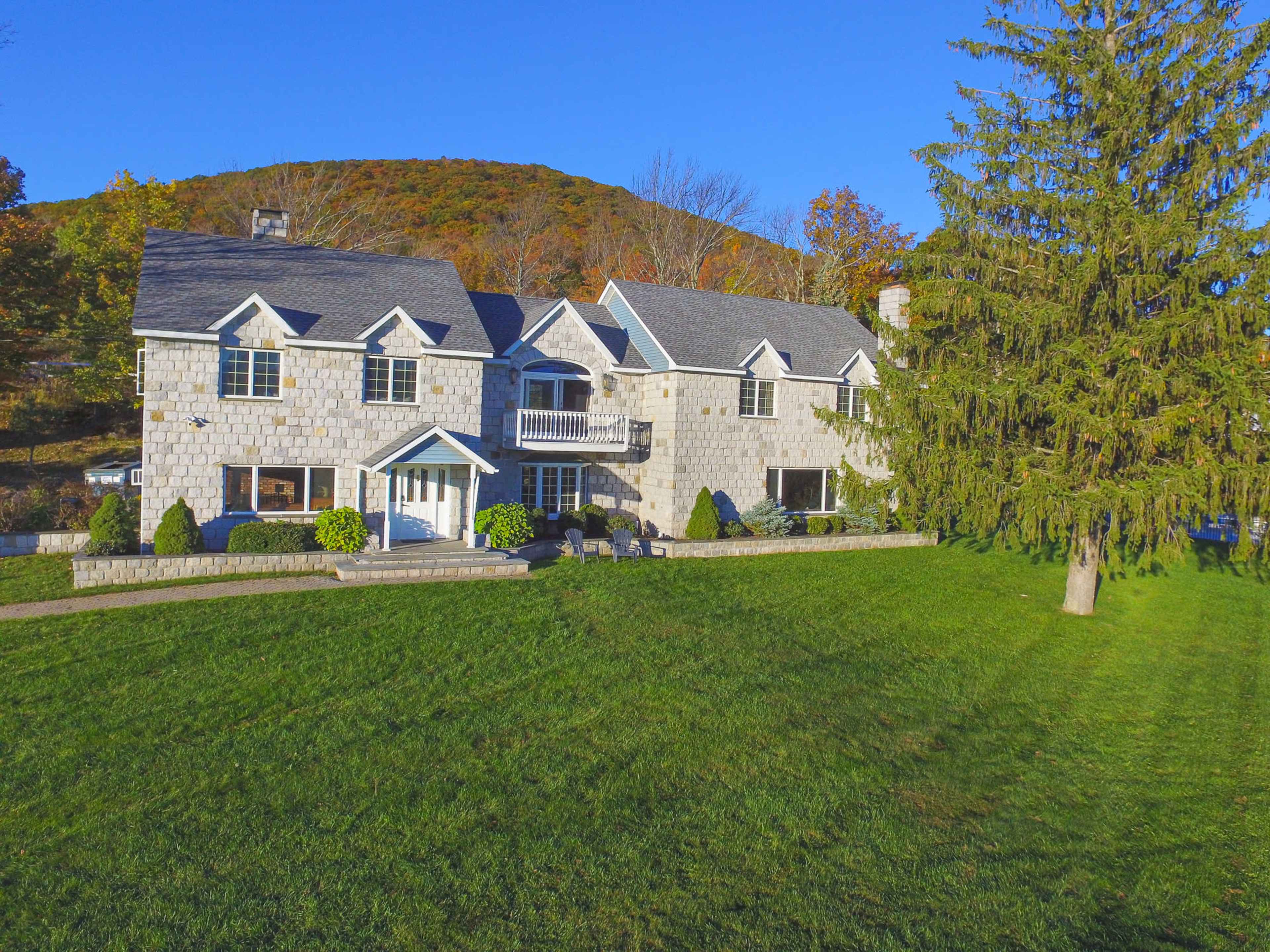 A large, stone house with multiple gables sits on a green lawn, backed by a hillside covered in autumn trees.