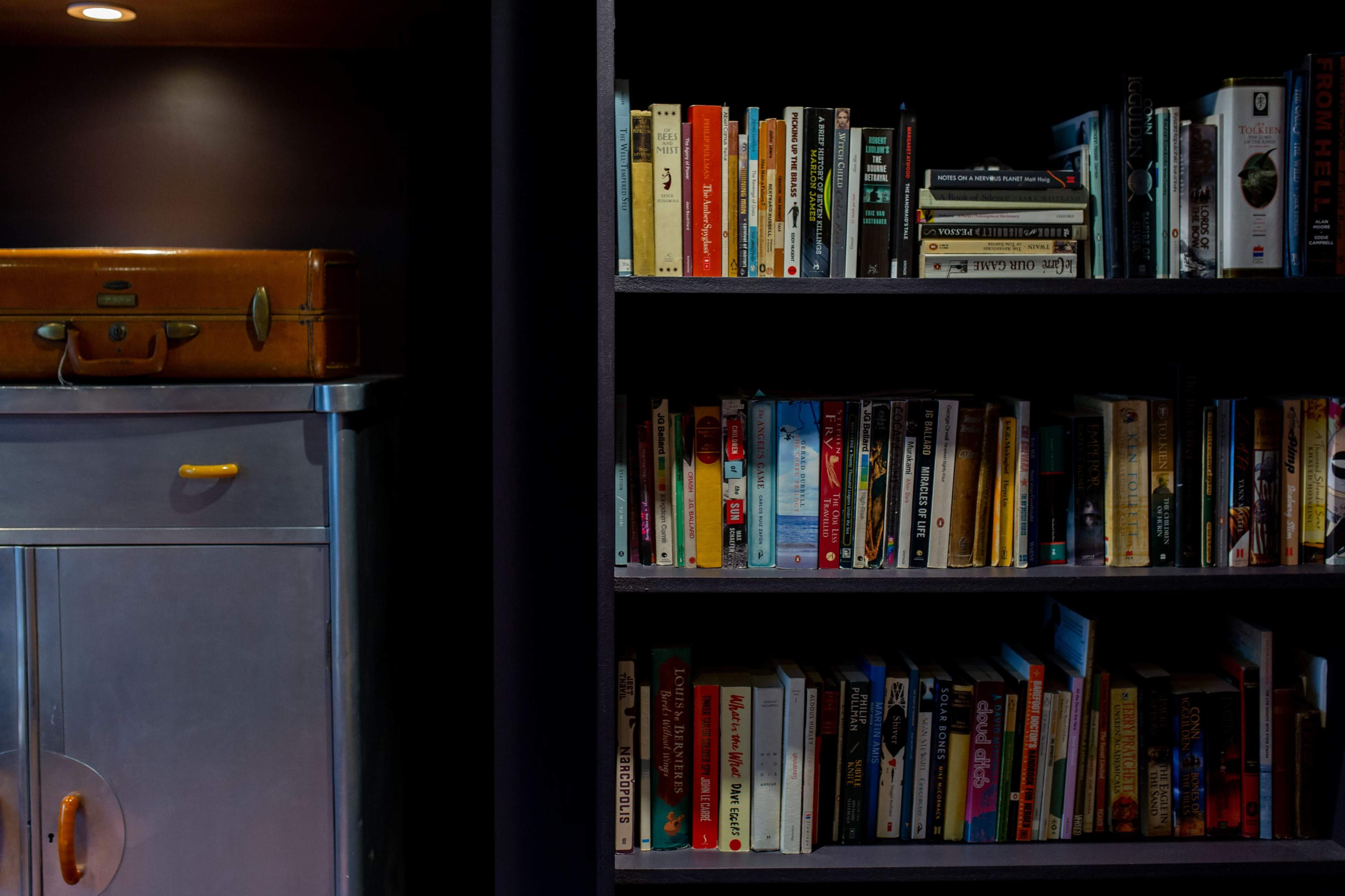 A wooden shelf filled with various books is positioned next to a metallic cabinet and a vintage suitcase.