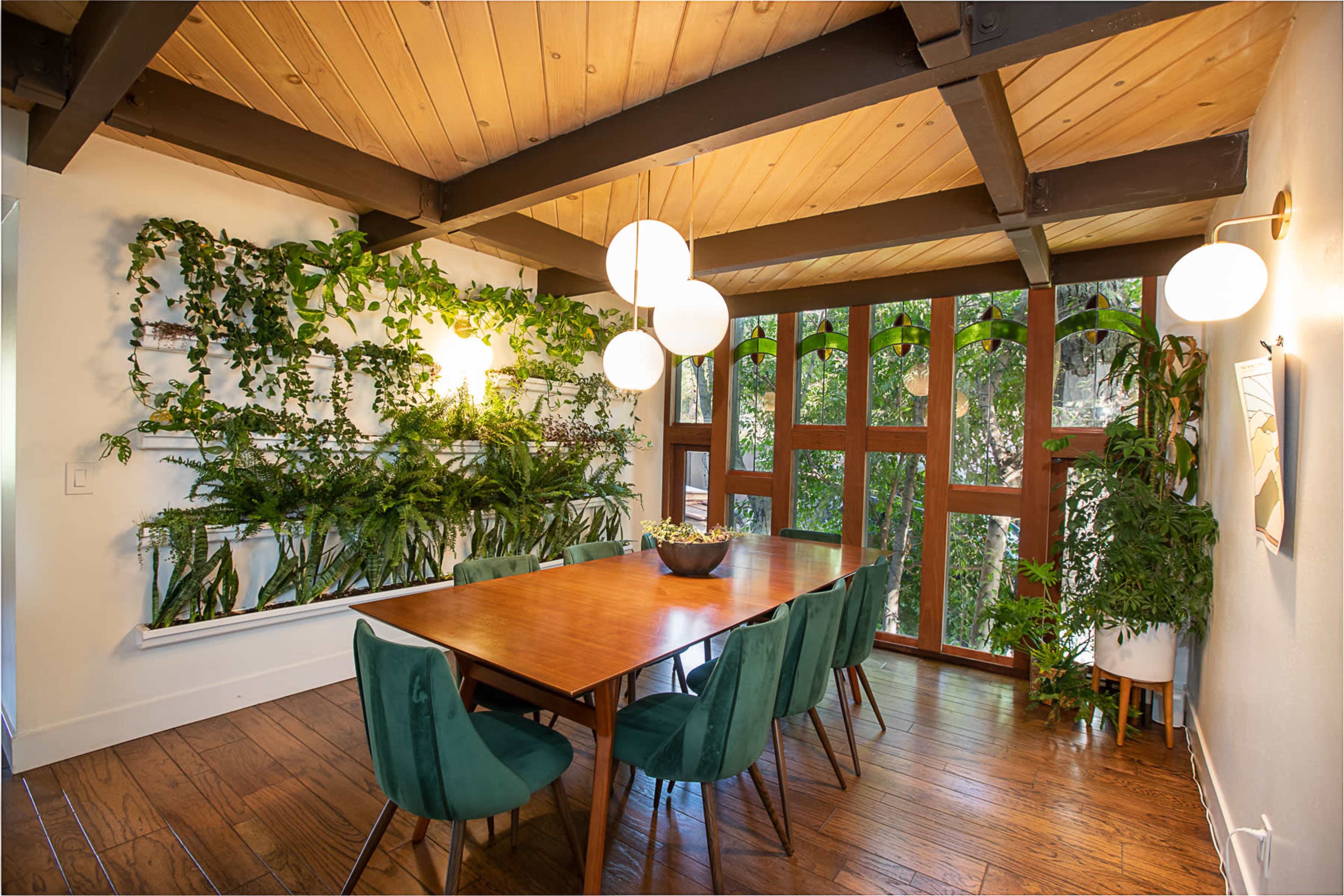 A dining room features a long wooden table surrounded by green chairs, with a lush vertical garden on one wall and large windows allowing natural light.