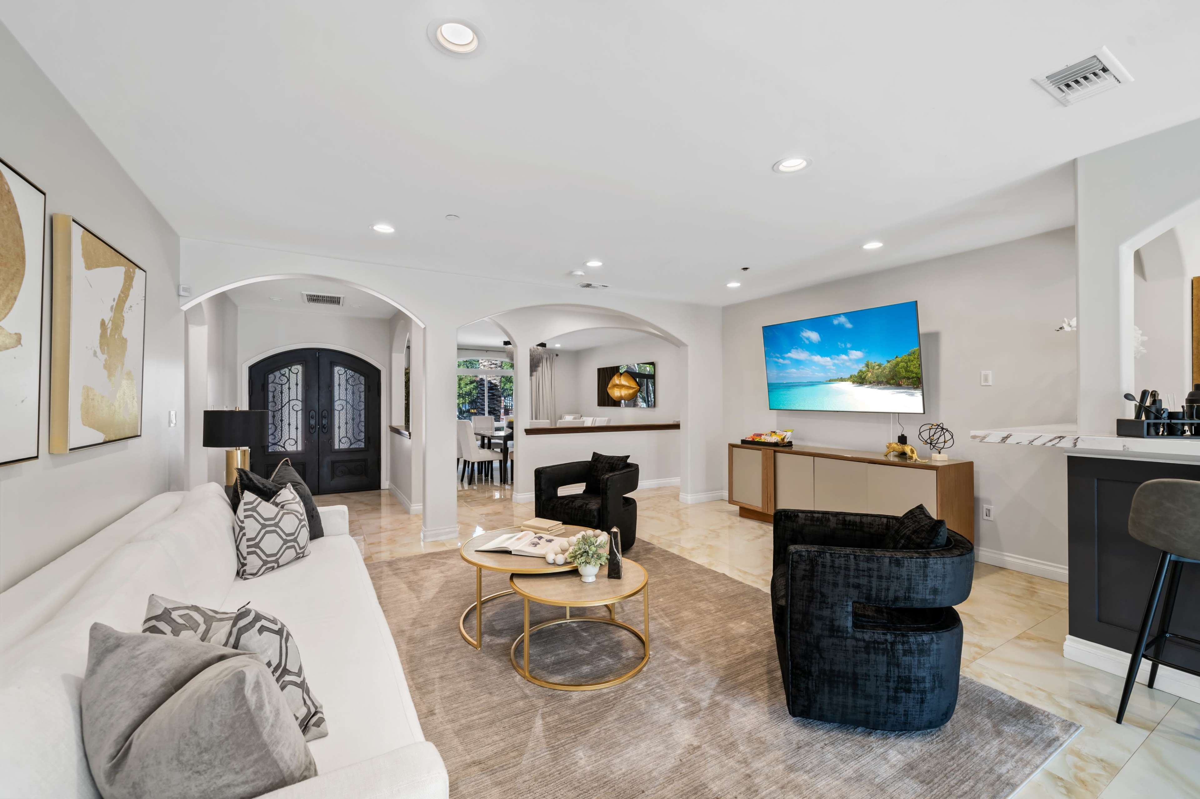 The living room features a white sofa with decorative pillows, a round coffee table, and a large TV displaying a beach scene, with archways leading to other areas of the home.