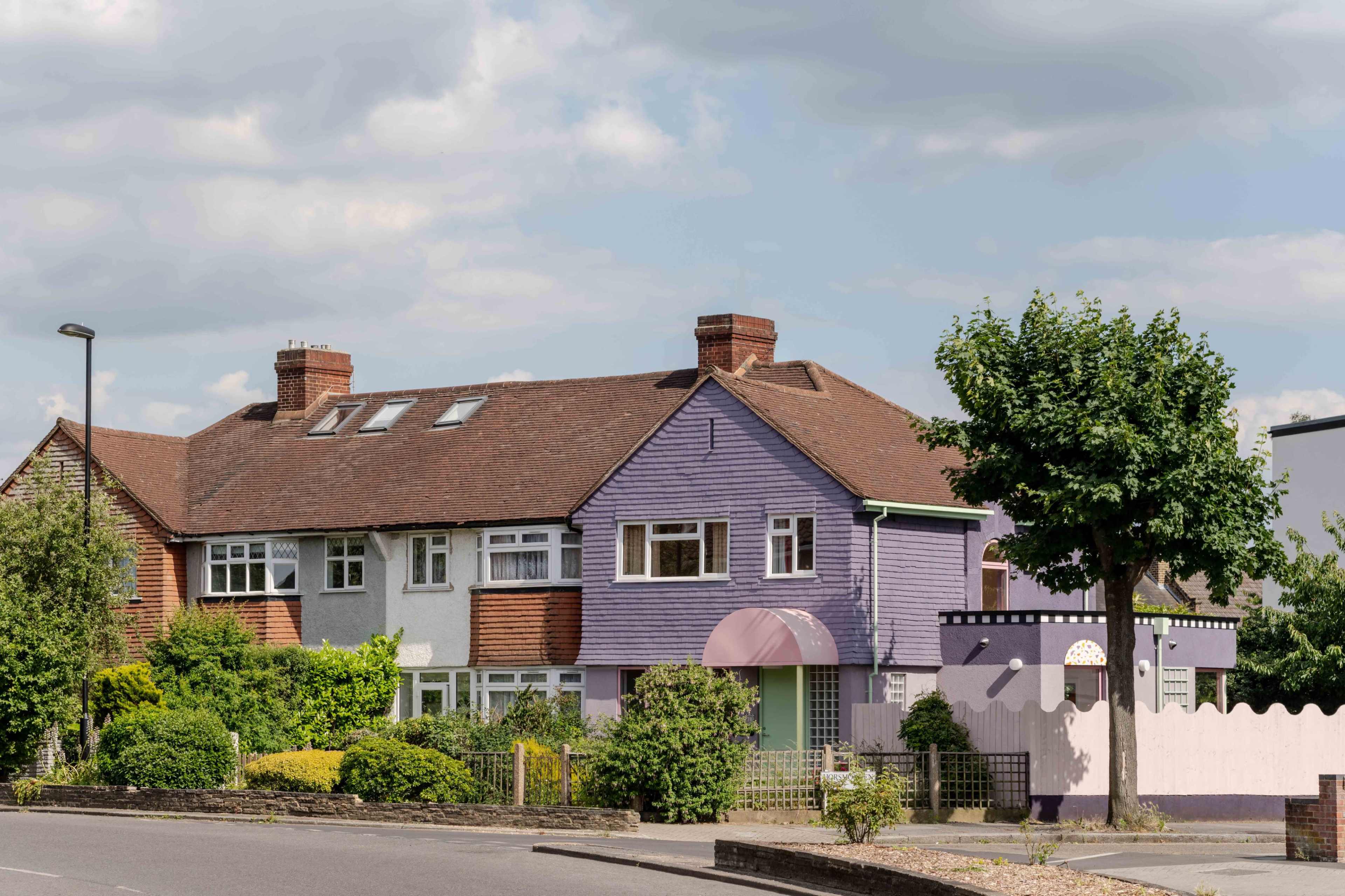 The image shows a two-story house with a purple exterior, a sloped roof, and a large tree in front, situated on a quiet street.
