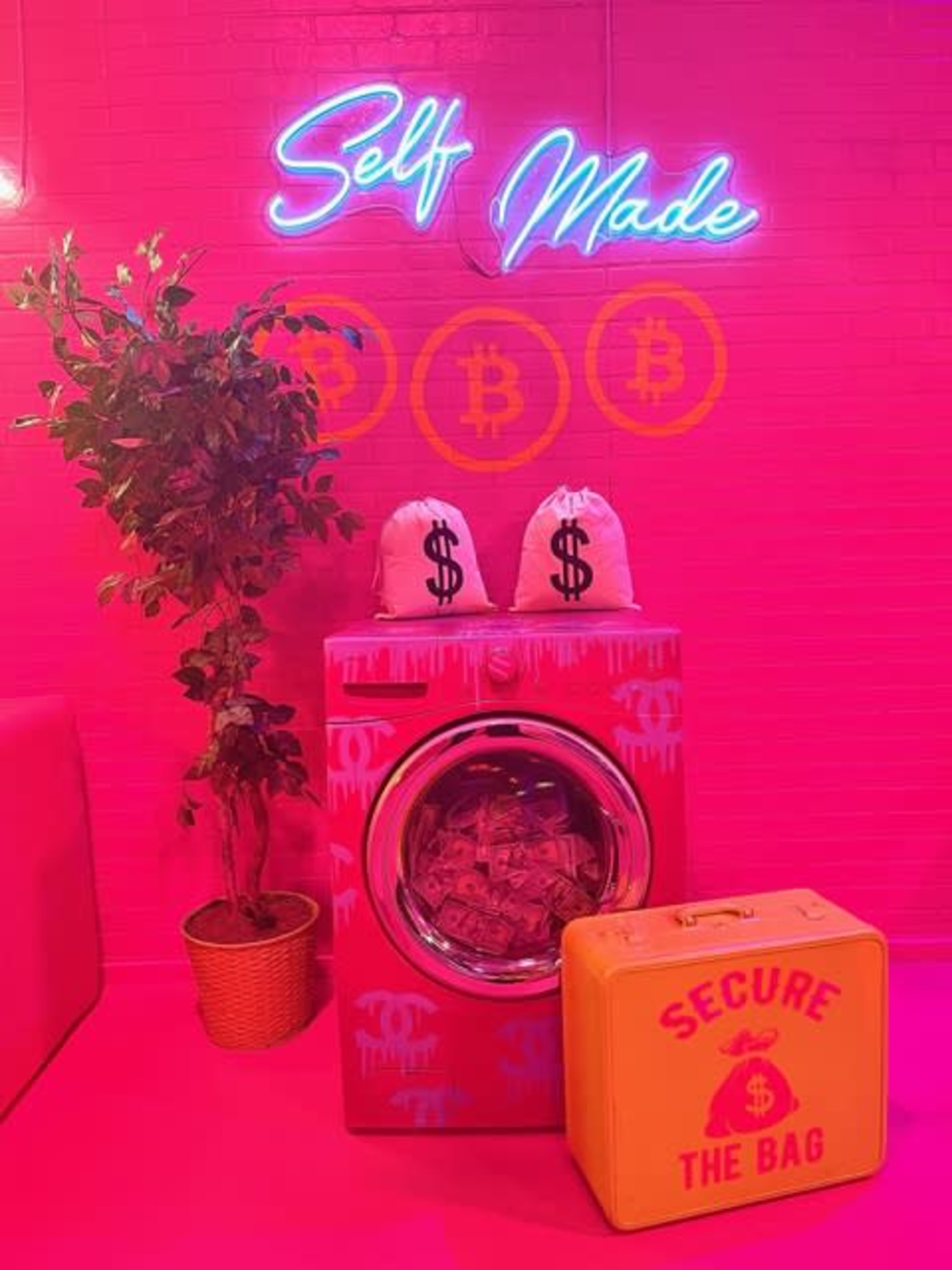 A vibrant pink room with a graffiti-style washing machine adorned with money and dollar-sign bags, alongside a bright yellow box labeled "SECURE THE BAG" and a neon sign reading "Self Made."
