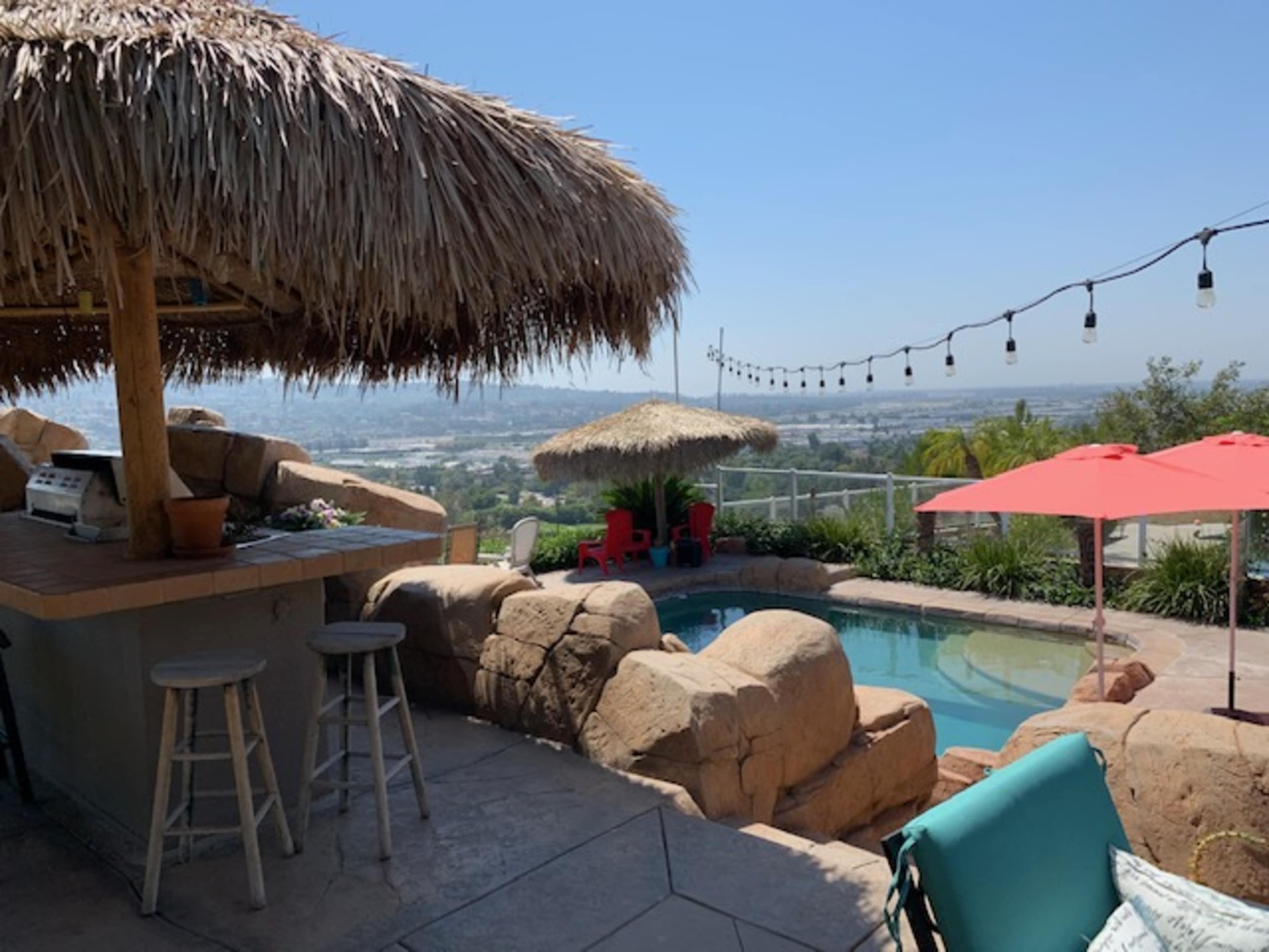 A pool area featuring a thatched gazebo, outdoor bar with stools, and seating surrounded by lush greenery and umbrellas.