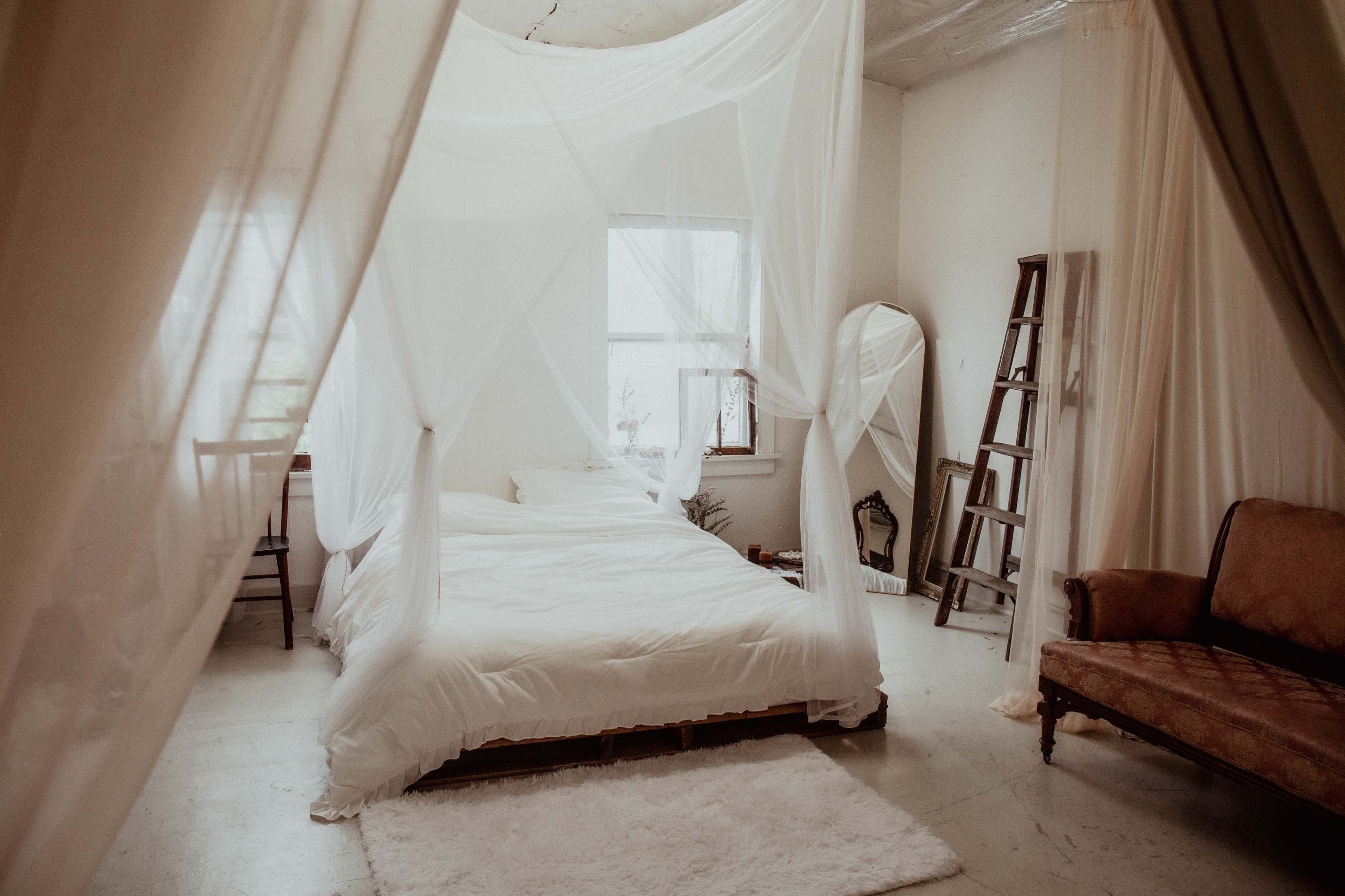 A bright bedroom features a large bed with white bedding draped in sheer canopies, accompanied by a small side chair, a ladder, and a vintage sofa along the walls.
