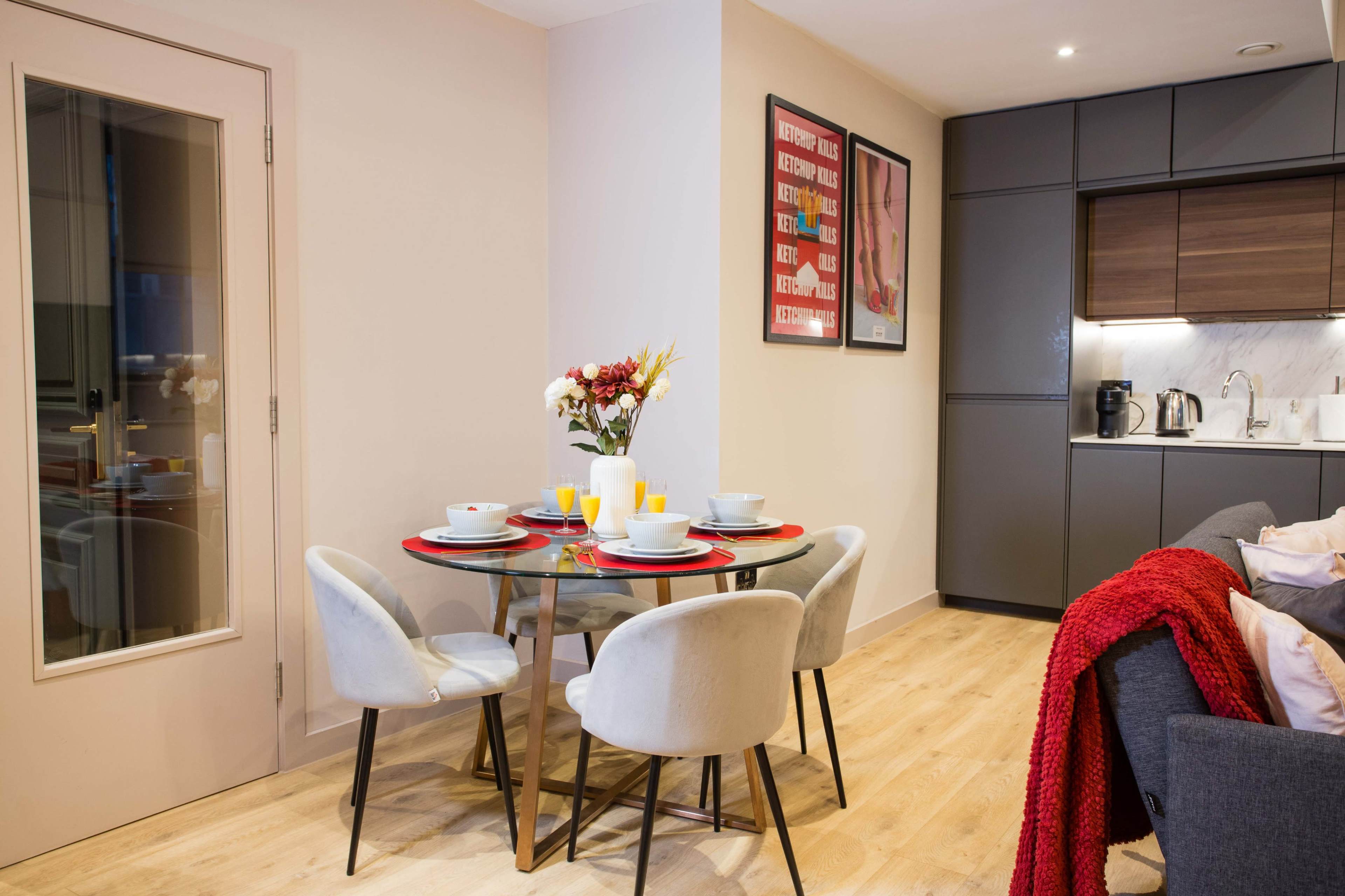 A modern dining area features a round glass table set with dishes and flowers, surrounded by four upholstered chairs, adjacent to a kitchen with sleek cabinetry.