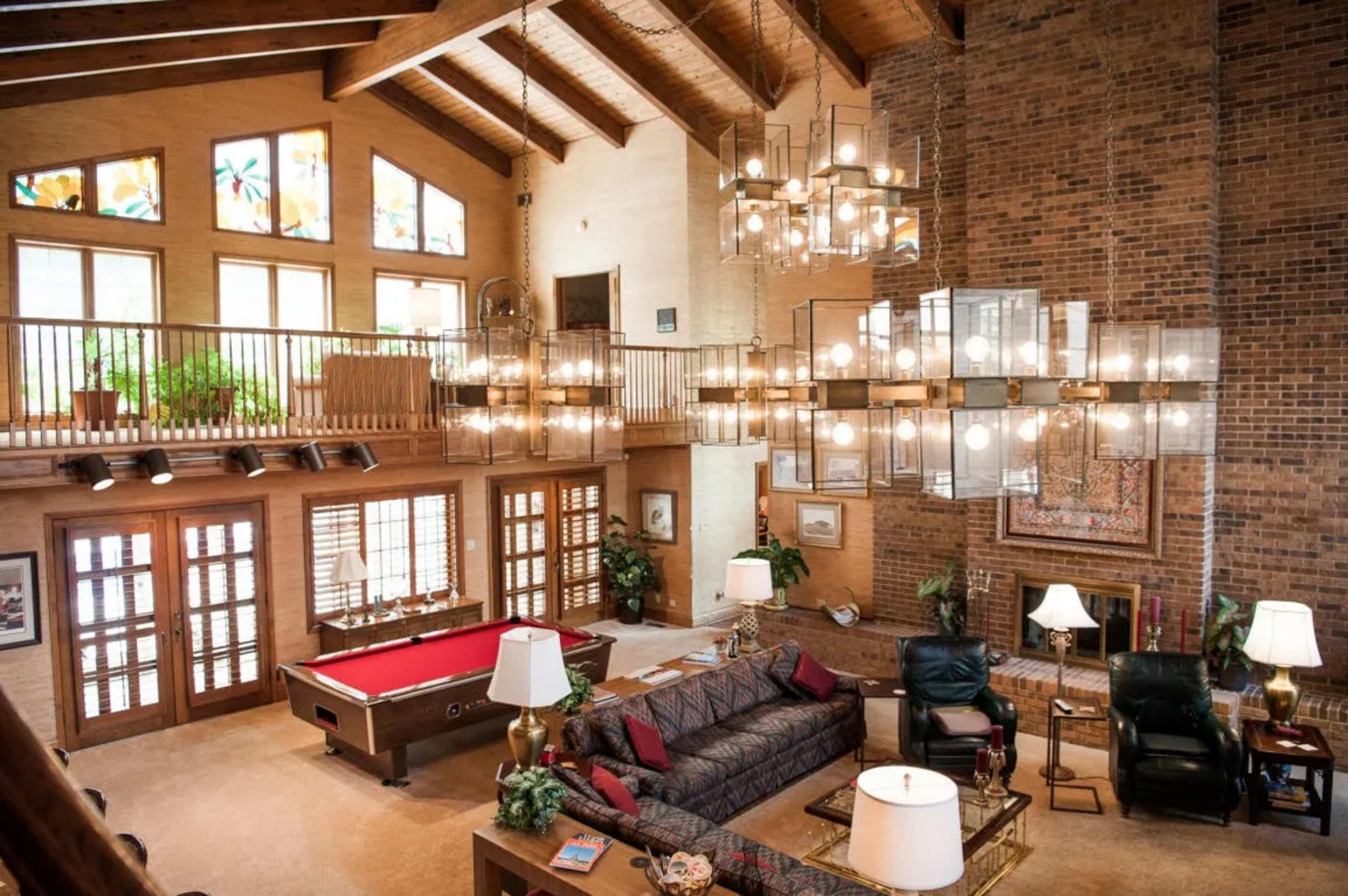 A spacious living room features tall, vaulted ceilings, large windows, and a pool table, with seating areas arranged around a central coffee table and illuminated by hanging chandeliers.