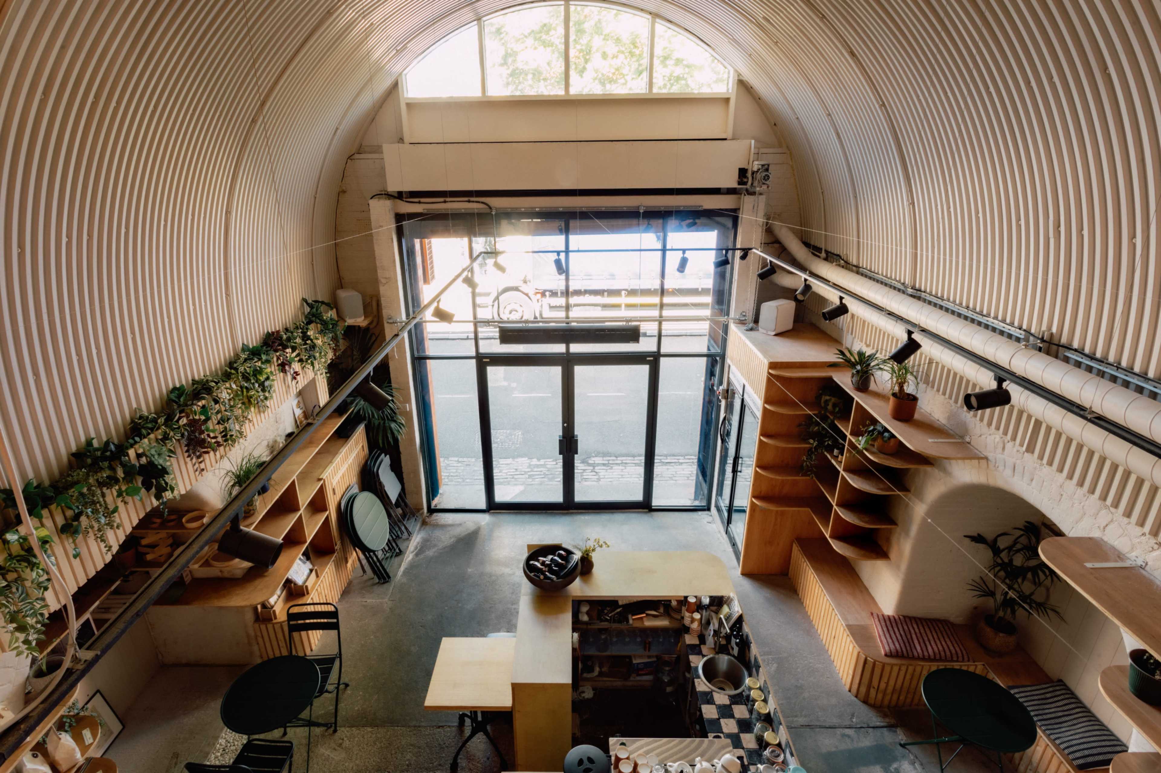 A modern café features an open layout with a large glass entrance, wooden shelves, and plants lining the walls.
