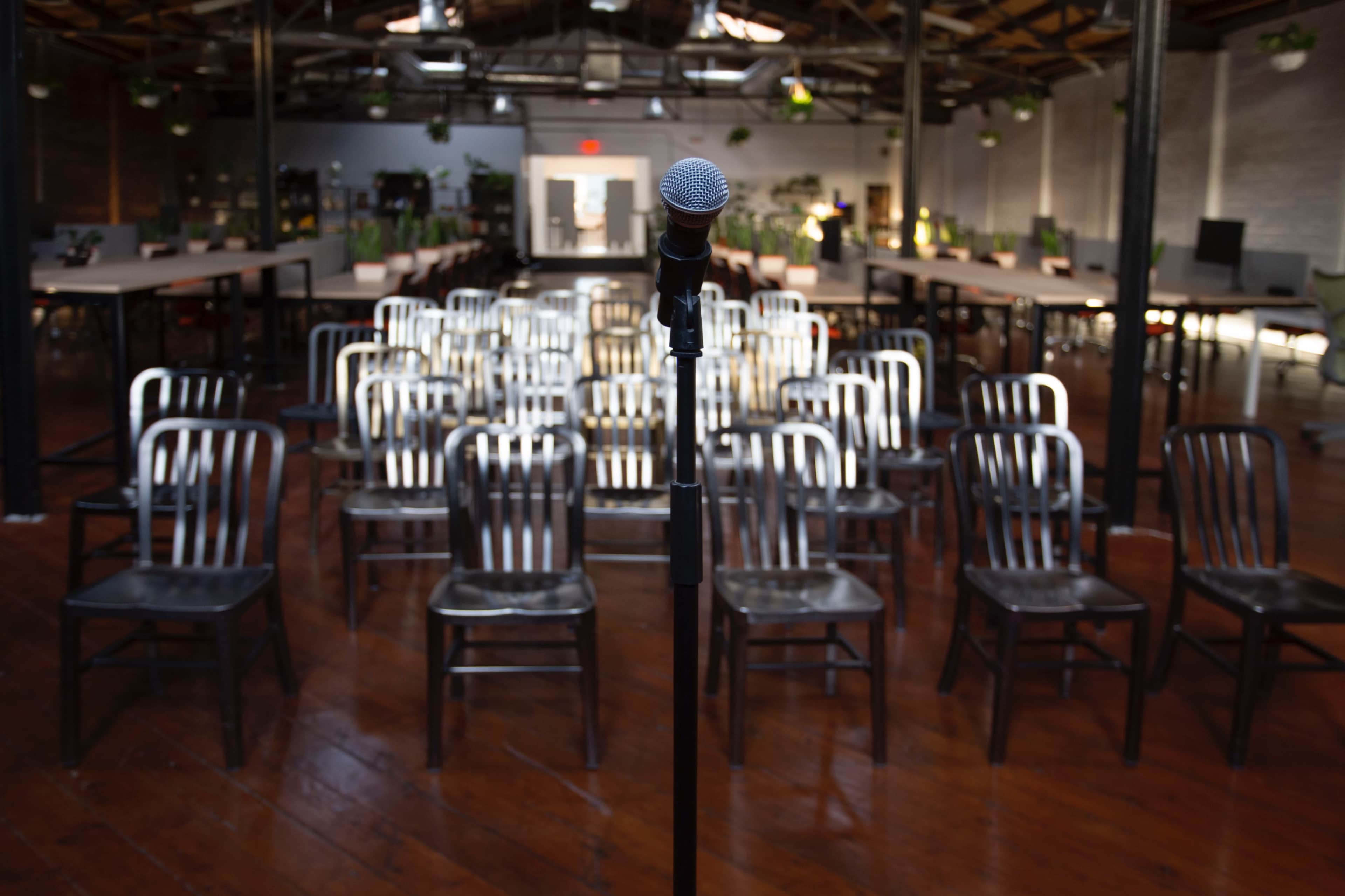 A microphone stands in the foreground facing a row of metal chairs arranged in an empty workspace.