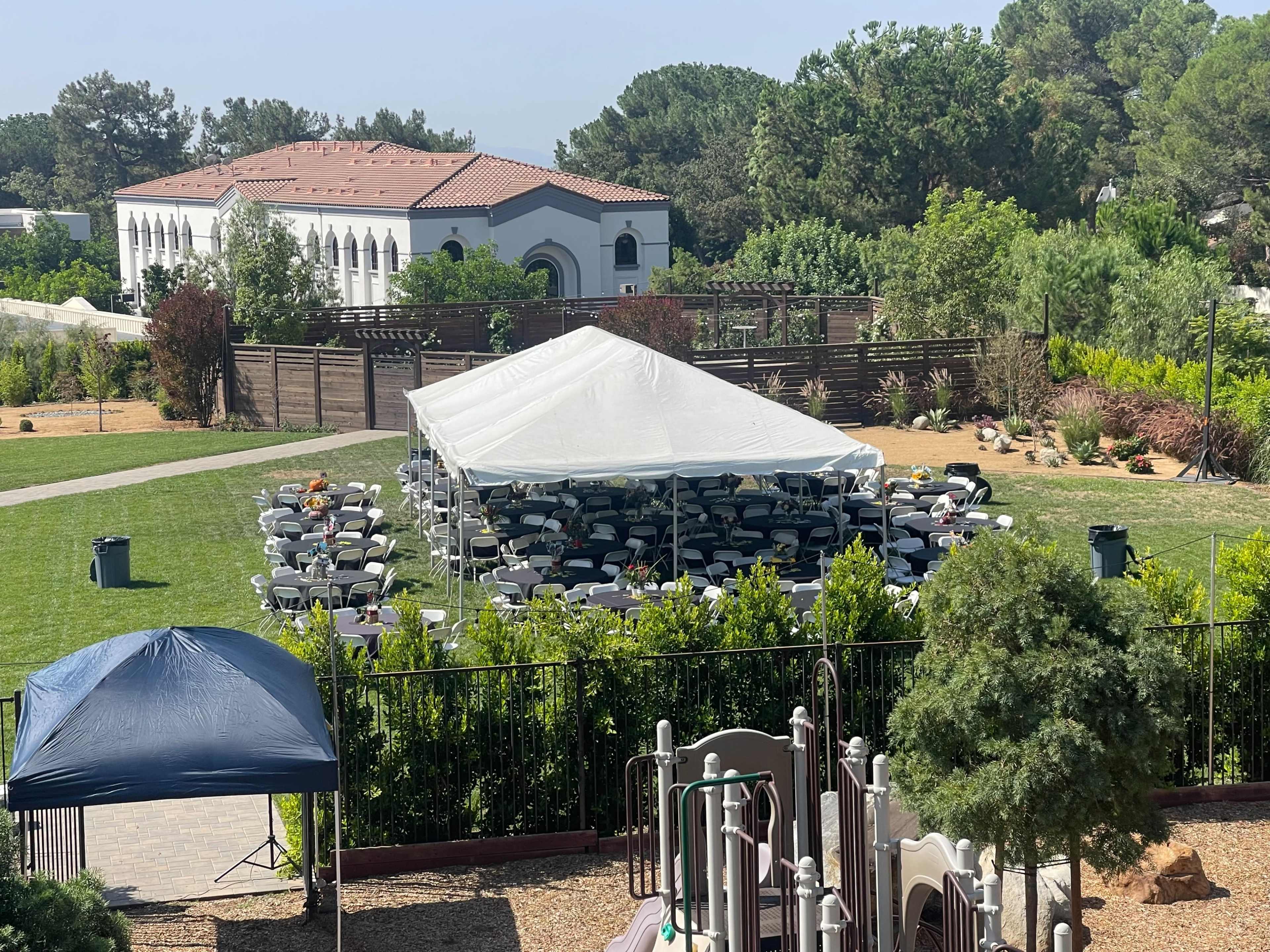 A large white tent filled with neatly arranged tables and chairs sits on a grassy area, with a playground in the foreground and a building visible in the background.