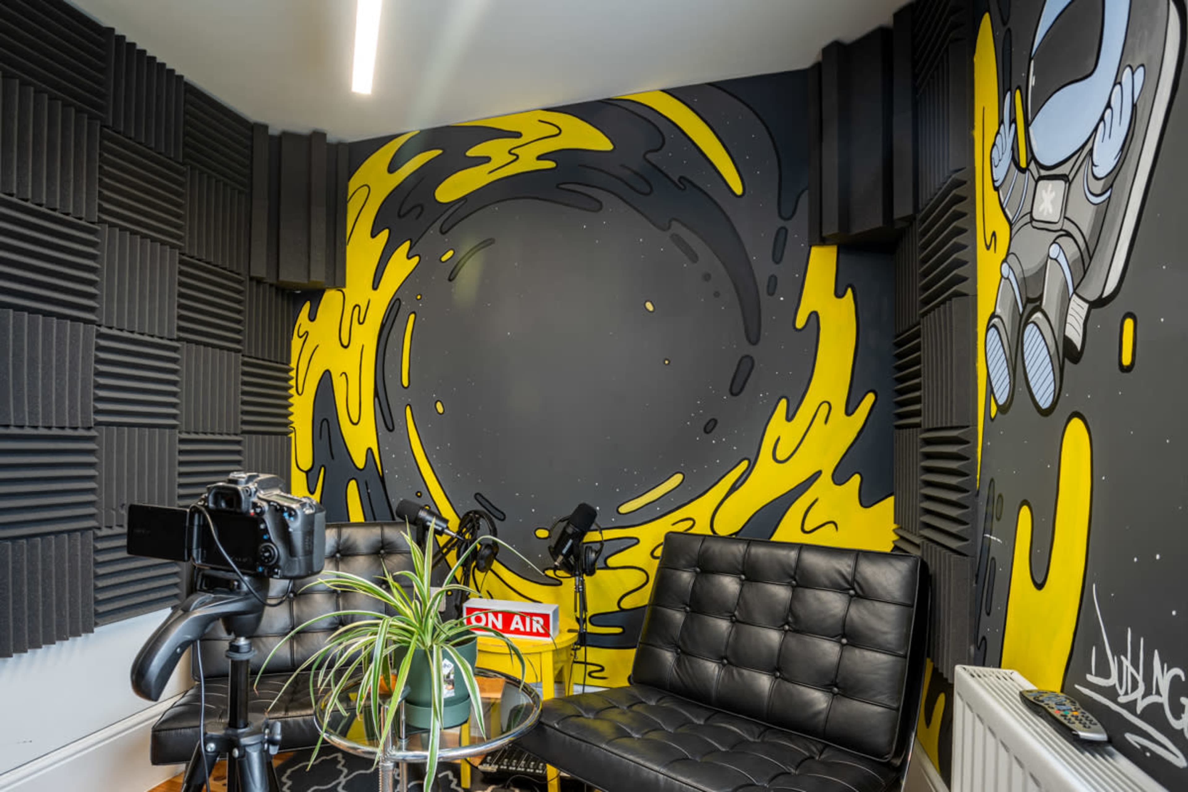 A corner of a recording studio featuring black soundproofing panels, a yellow and black mural, and seating arrangements with plants and recording equipment.
