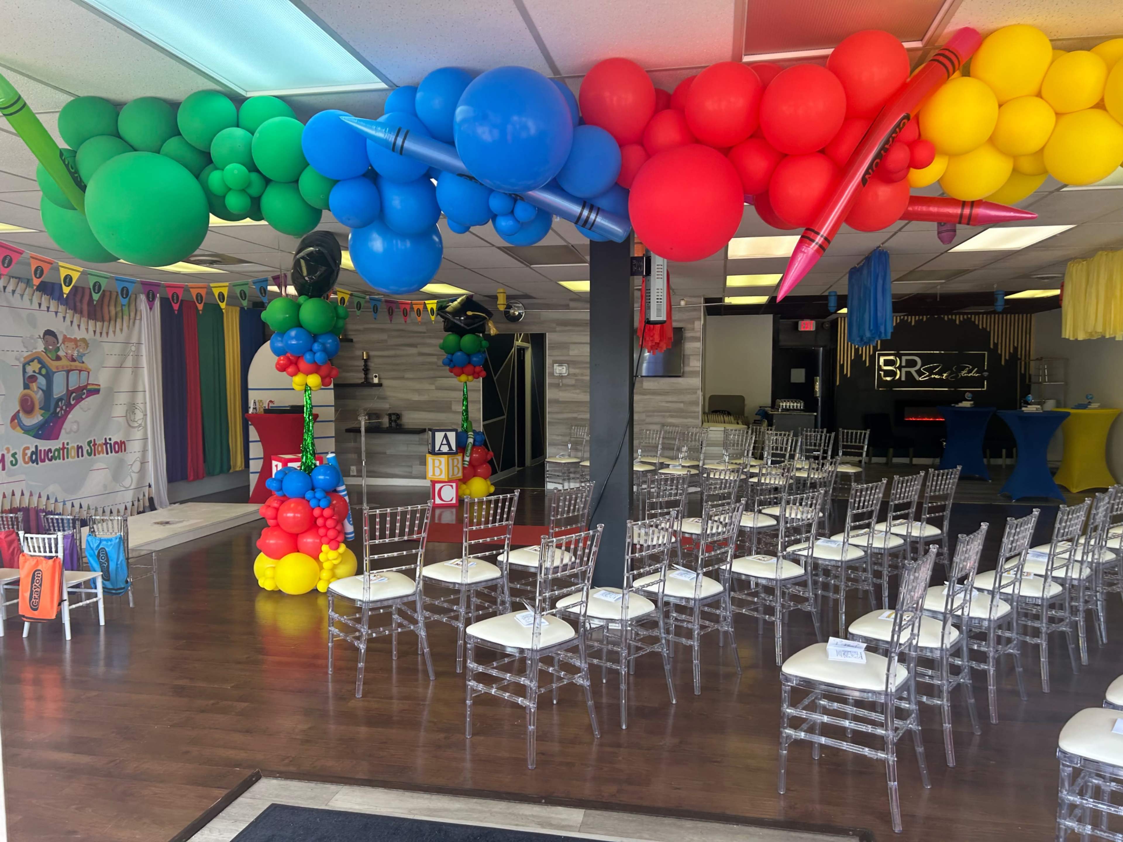 The image shows a colorful event space decorated with balloon arches and rows of clear chairs set up for an event.