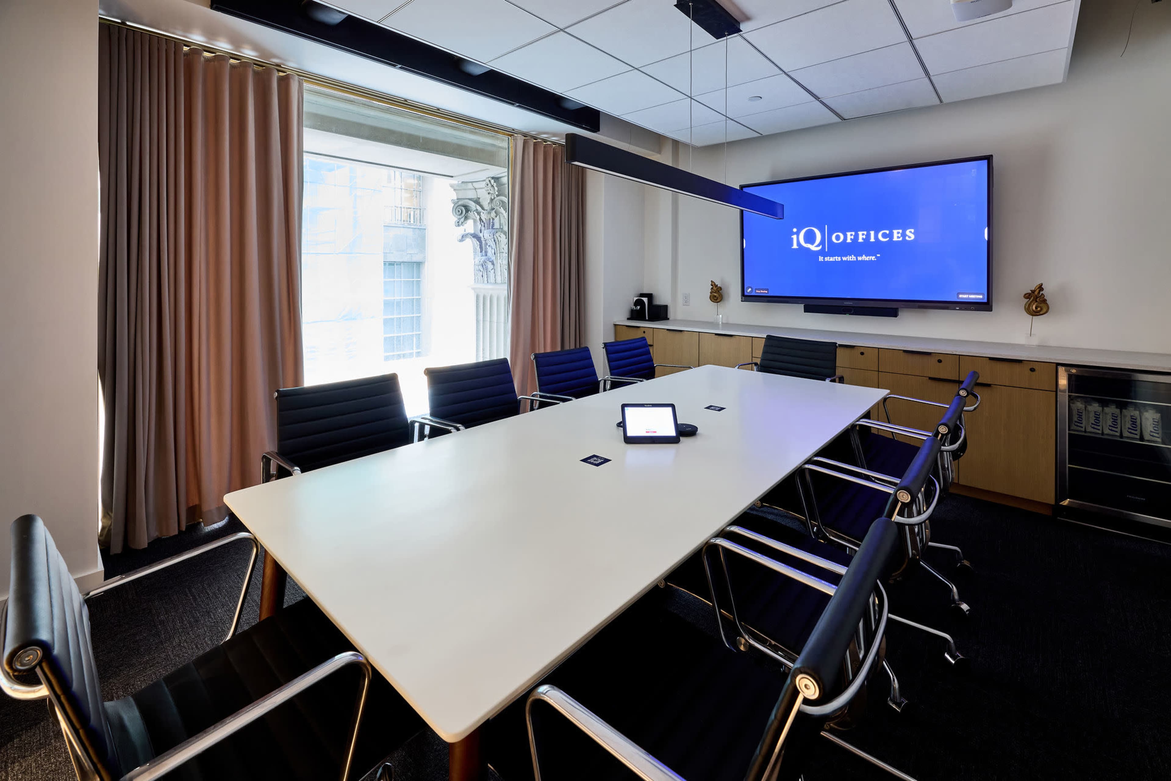 A modern conference room features a long white table surrounded by black chairs, with a large screen displaying the logo of IQ Offices on the wall.