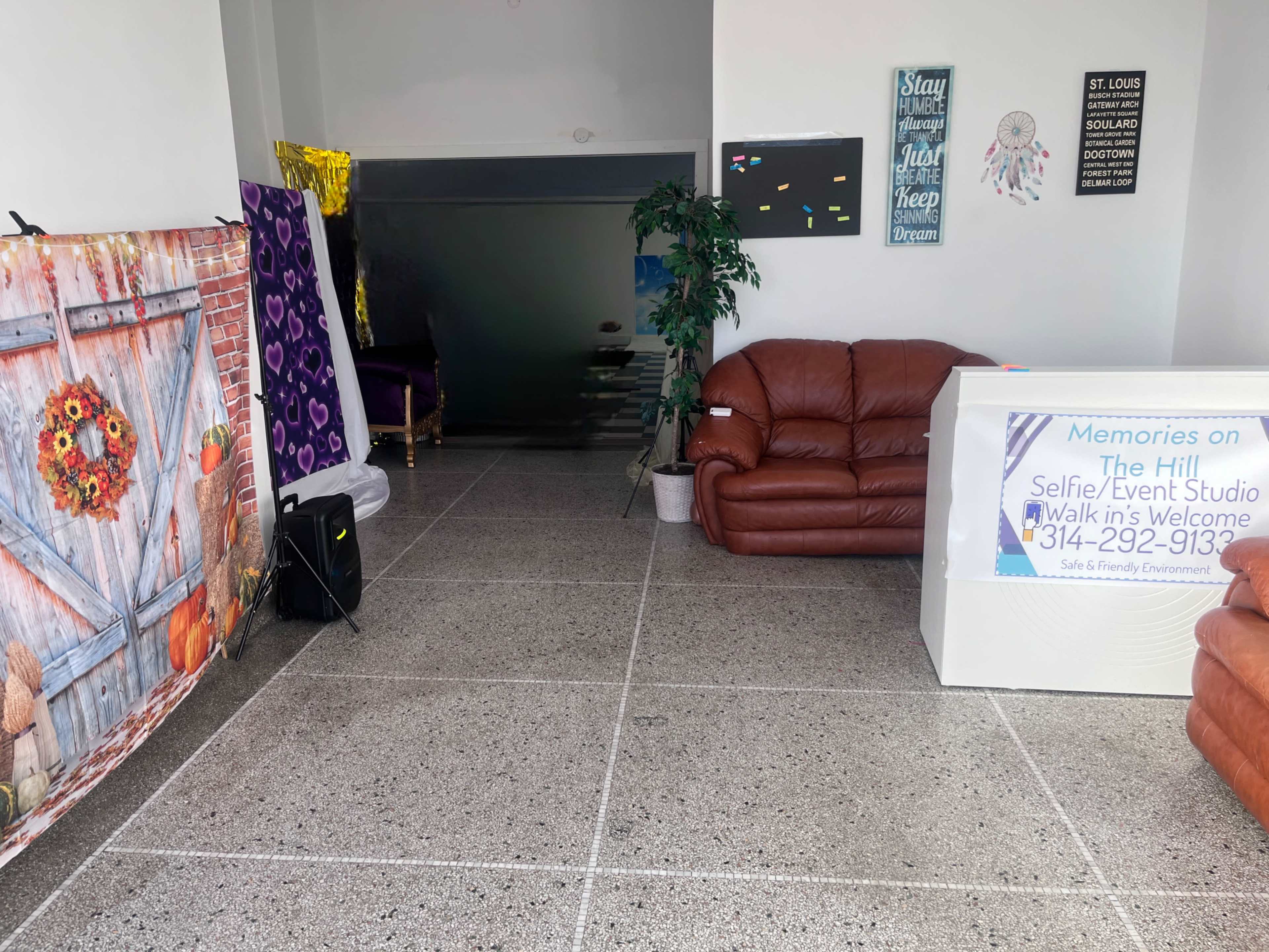 The image shows a bright, spacious room featuring two brown couches, a decorative backdrop with flowers, and a sign for a selfie/event studio.