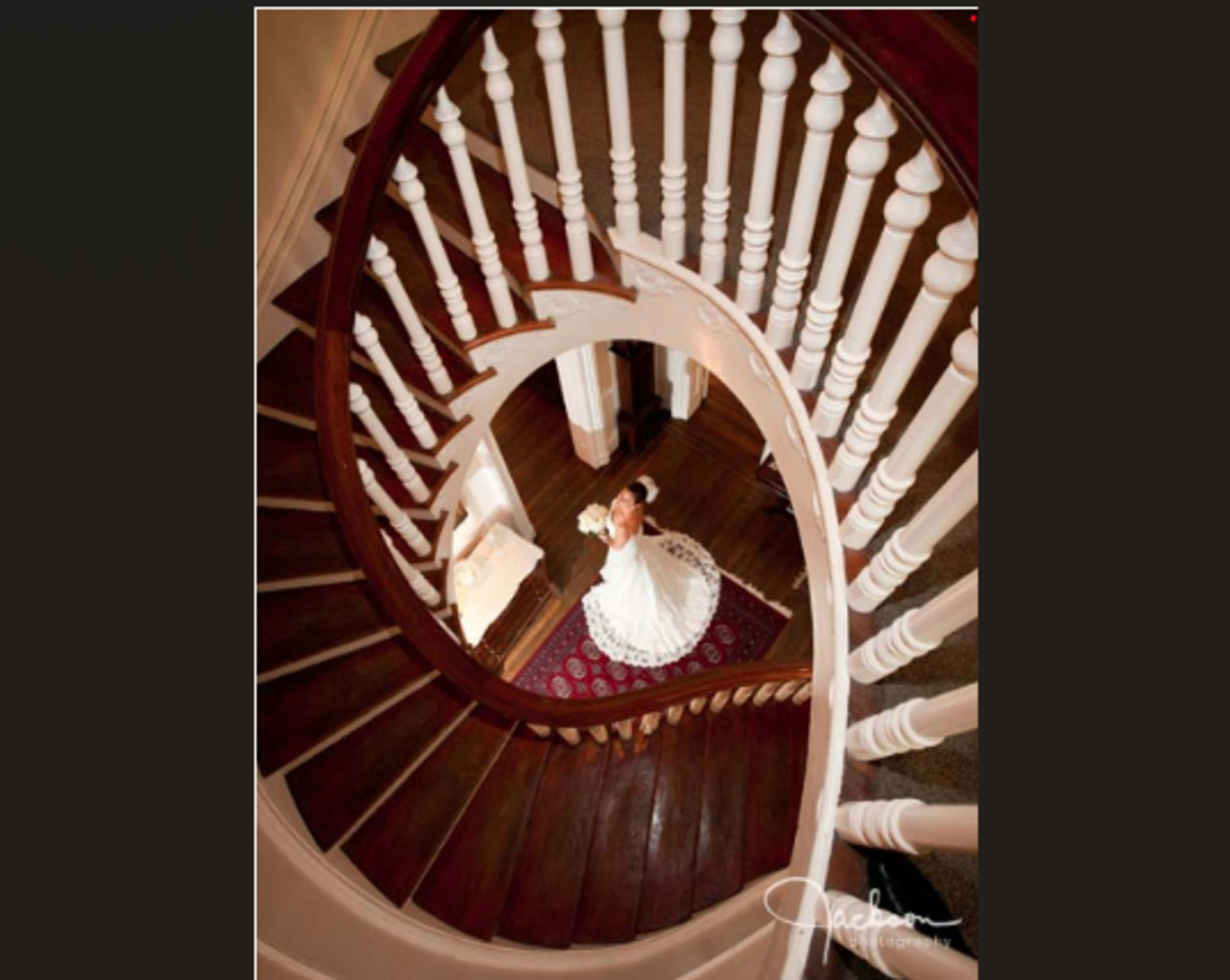 A bride in a white wedding dress stands at the base of a spiral staircase, holding a bouquet.
