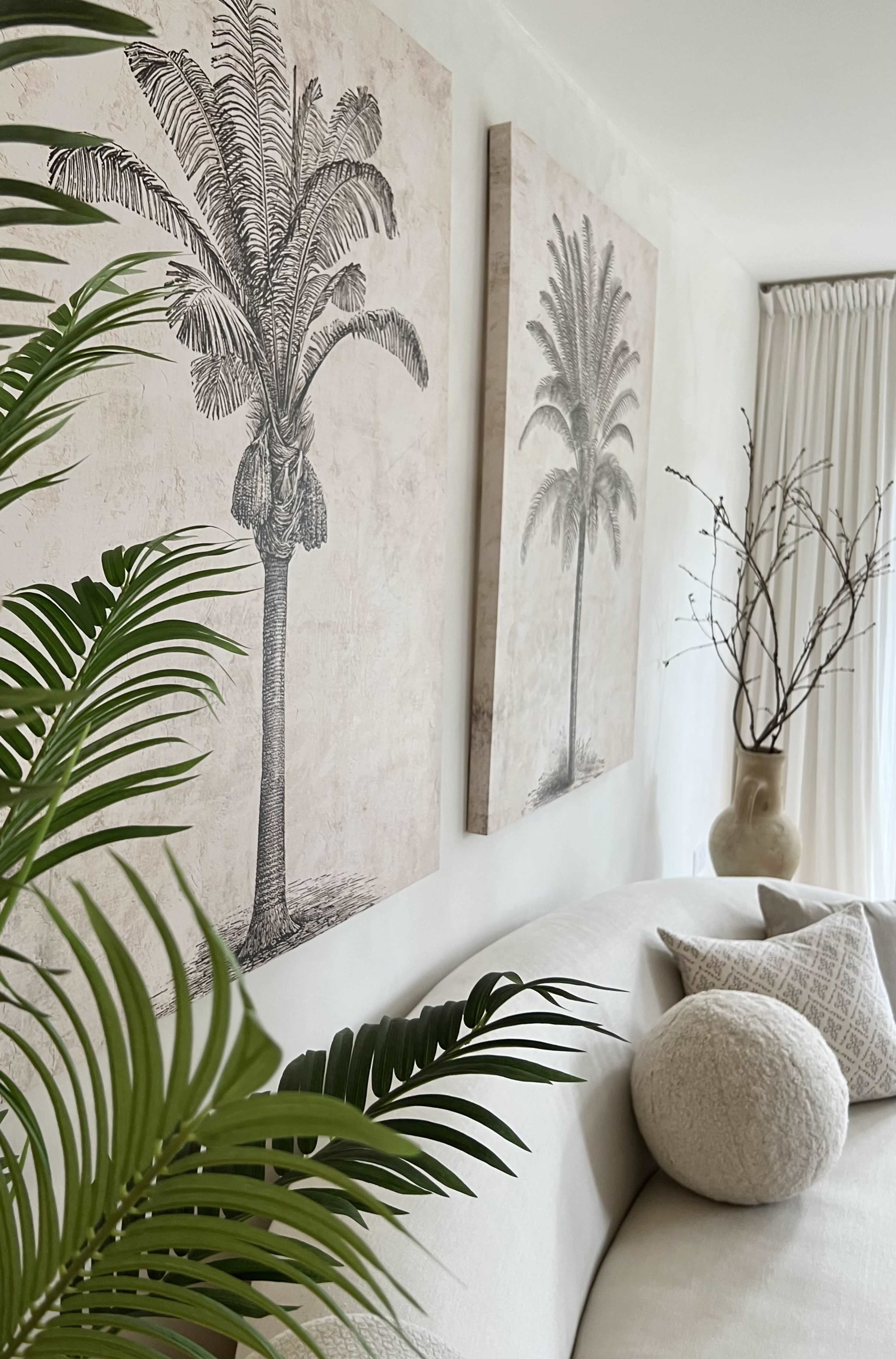 A beige sofa is positioned against a wall adorned with two prints of palm trees, accompanied by green leaves and a decorative vase.