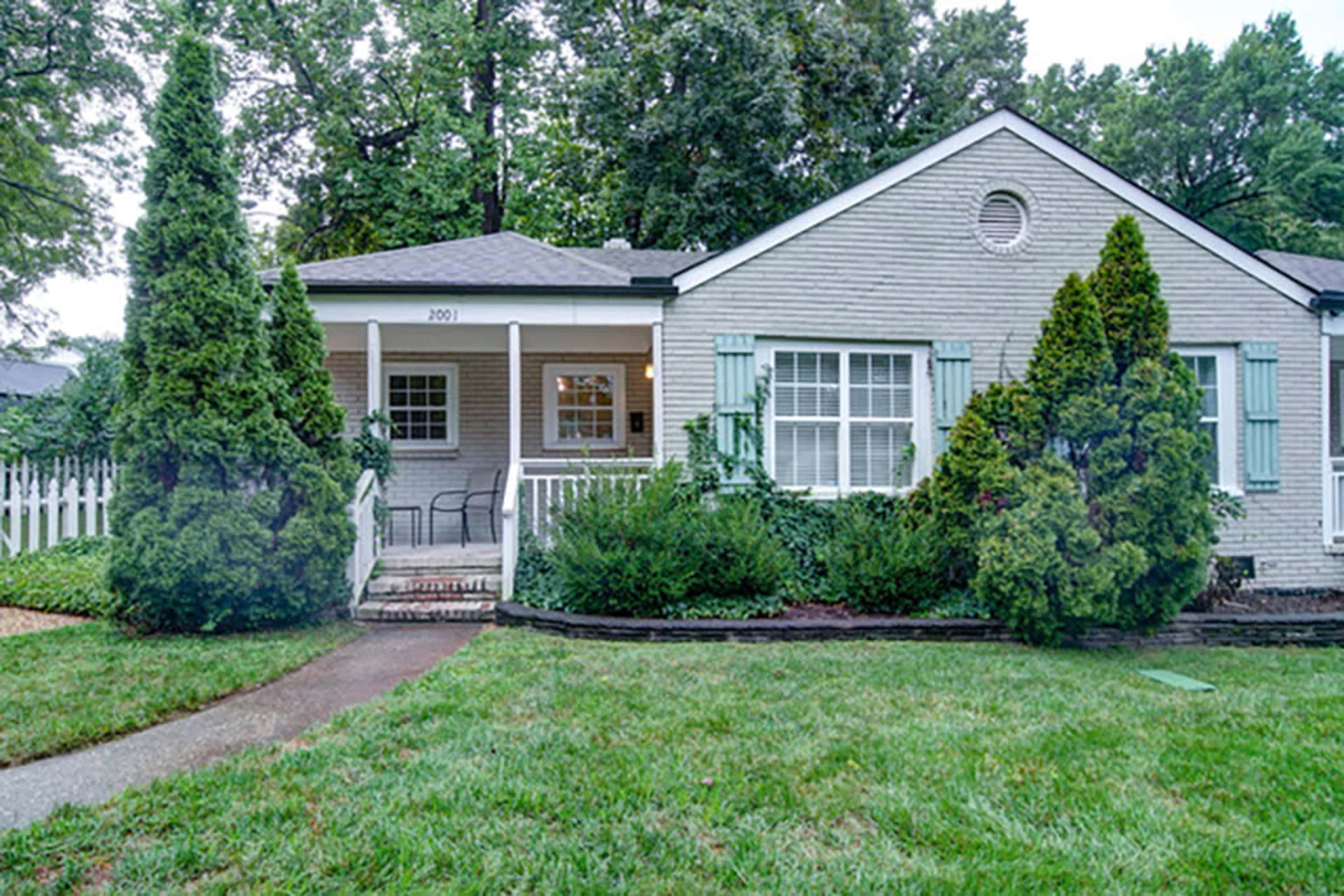 The image shows a single-story gray house with green shutters, a front porch, and a landscaped yard featuring shrubs and a pathway.