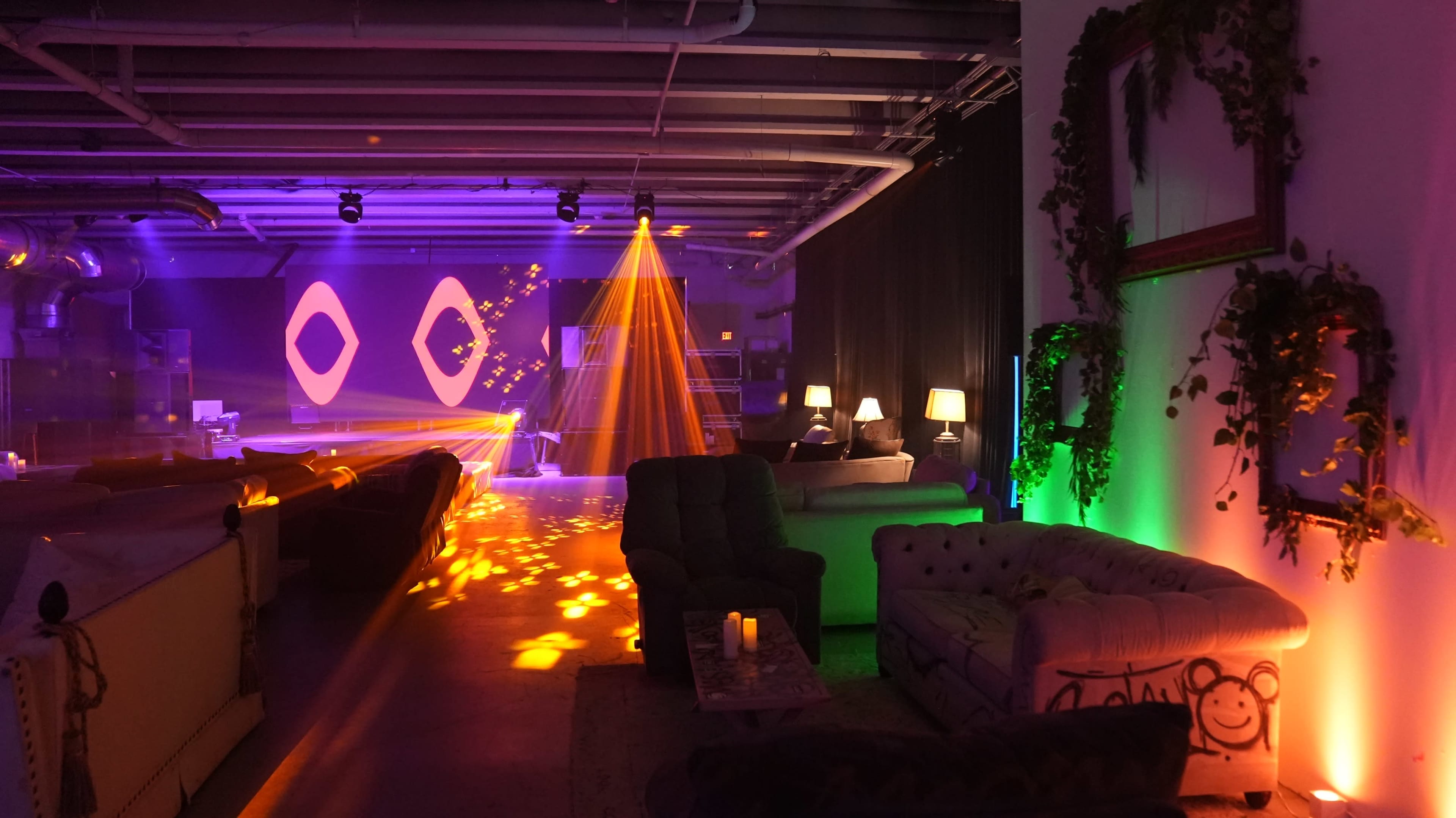 The image shows a dimly lit lounge area with colorful lighting, featuring sofas, lamps, and a stage backdrop with geometric designs.