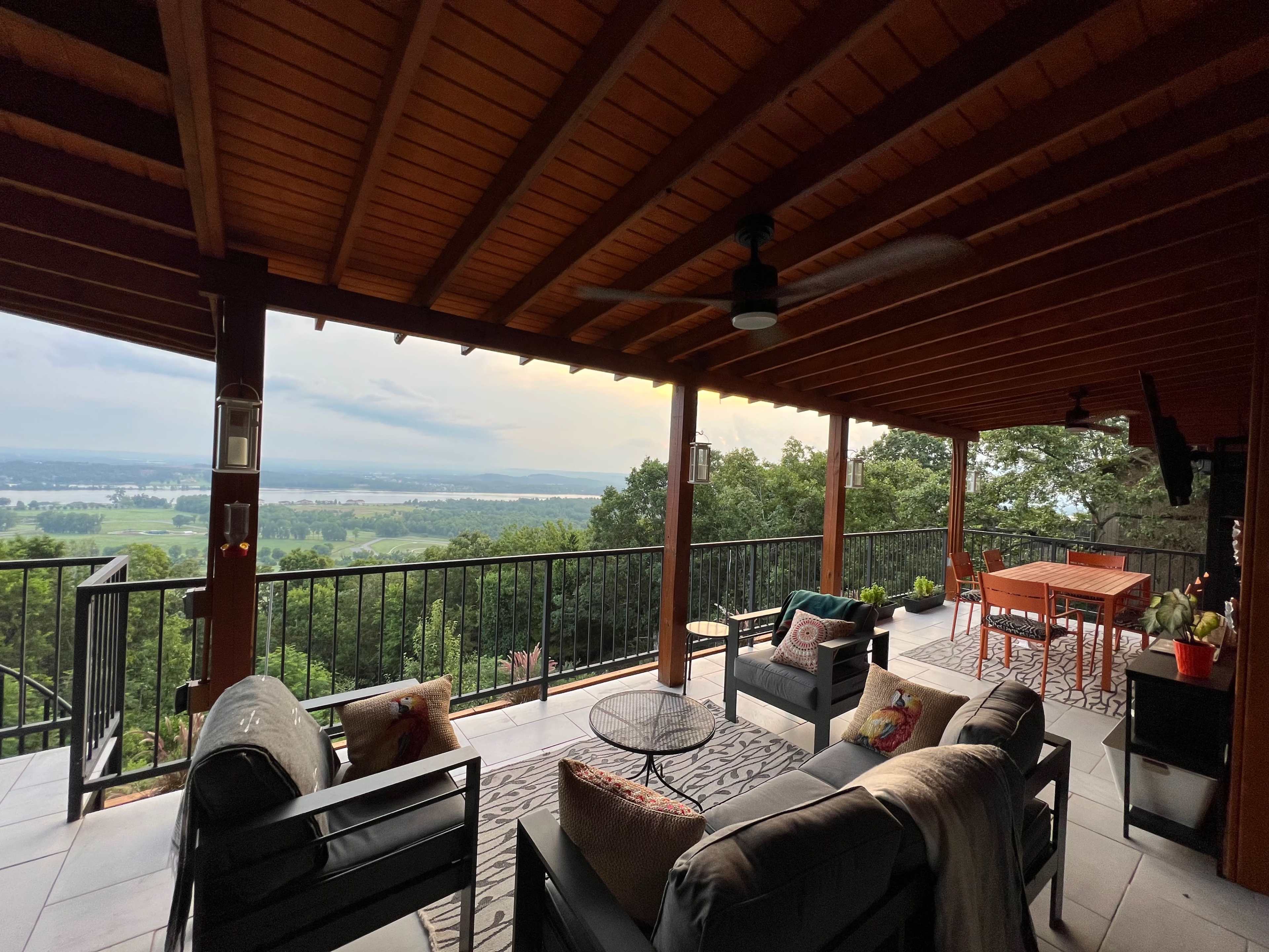 A covered patio with seating and a table, overlooking a green landscape under a cloudy sky.