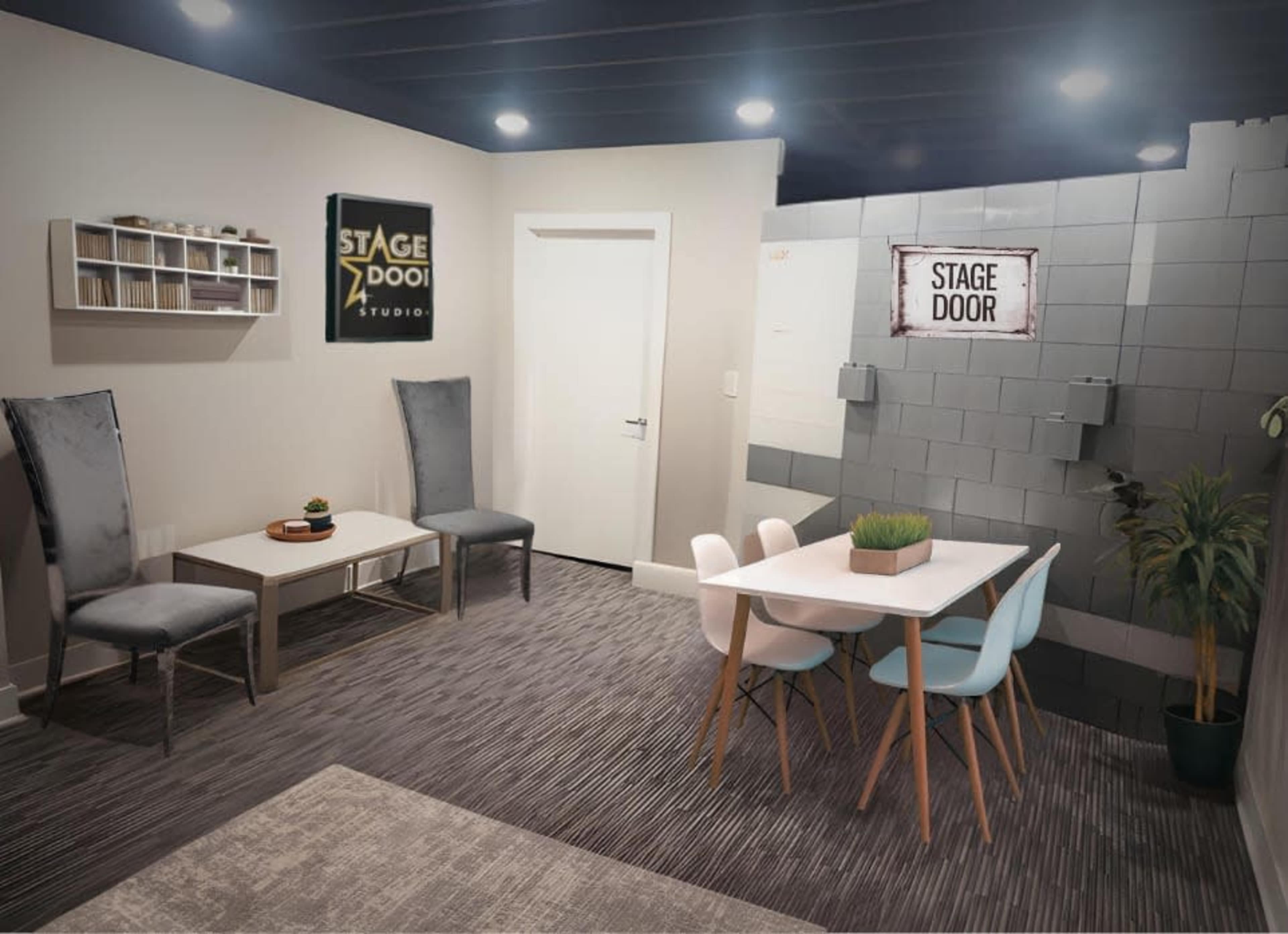 The image shows a modern studio or rehearsal space with a seating area featuring two chairs and a coffee table, alongside a dining table with colorful chairs, a plant centerpiece, and a wall sign that reads "STAGE DOOR."
