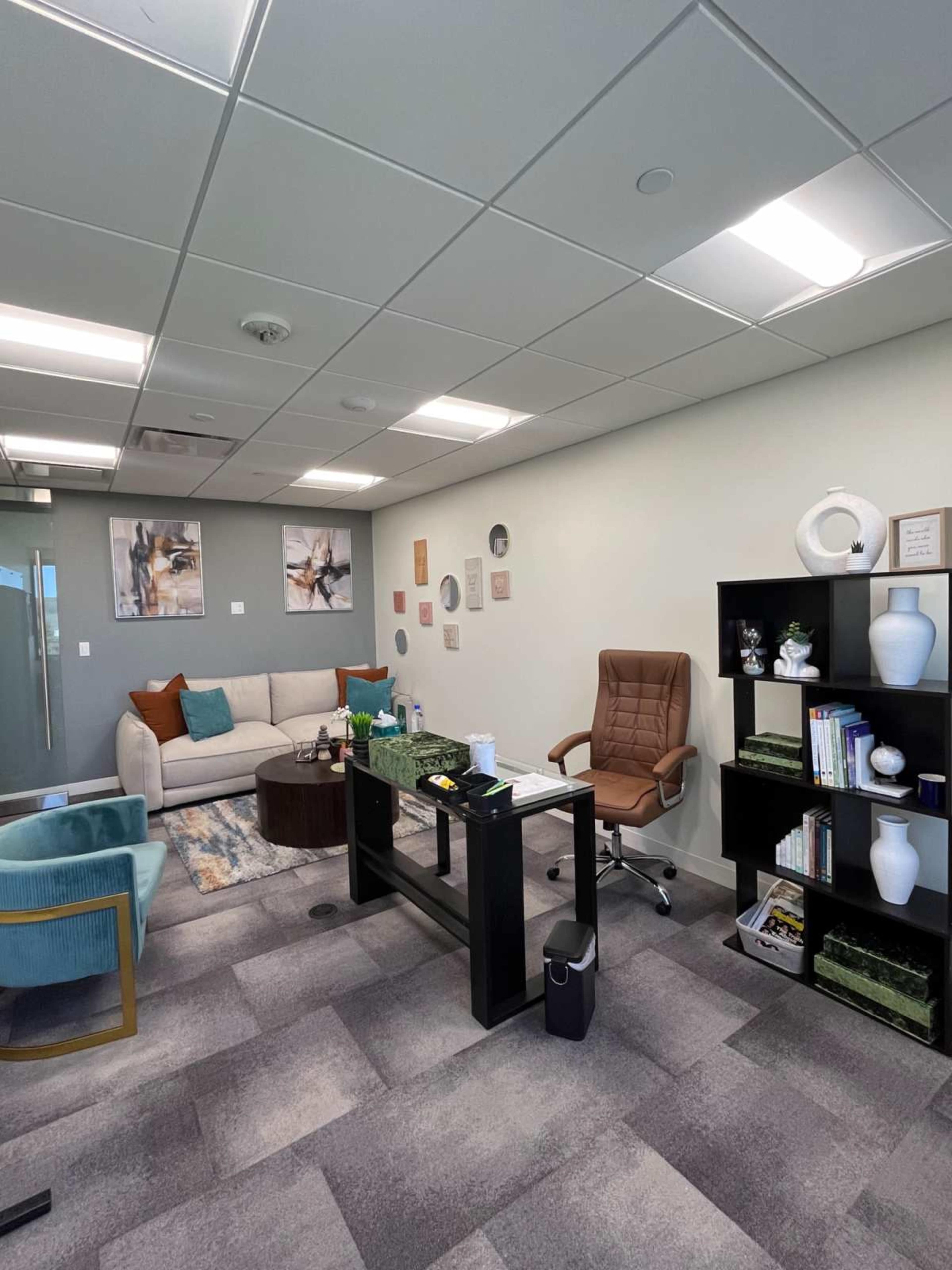 A modern office space featuring a beige sofa, a brown ergonomic chair, a black desk with green plants, and a bookshelf with decorative items.