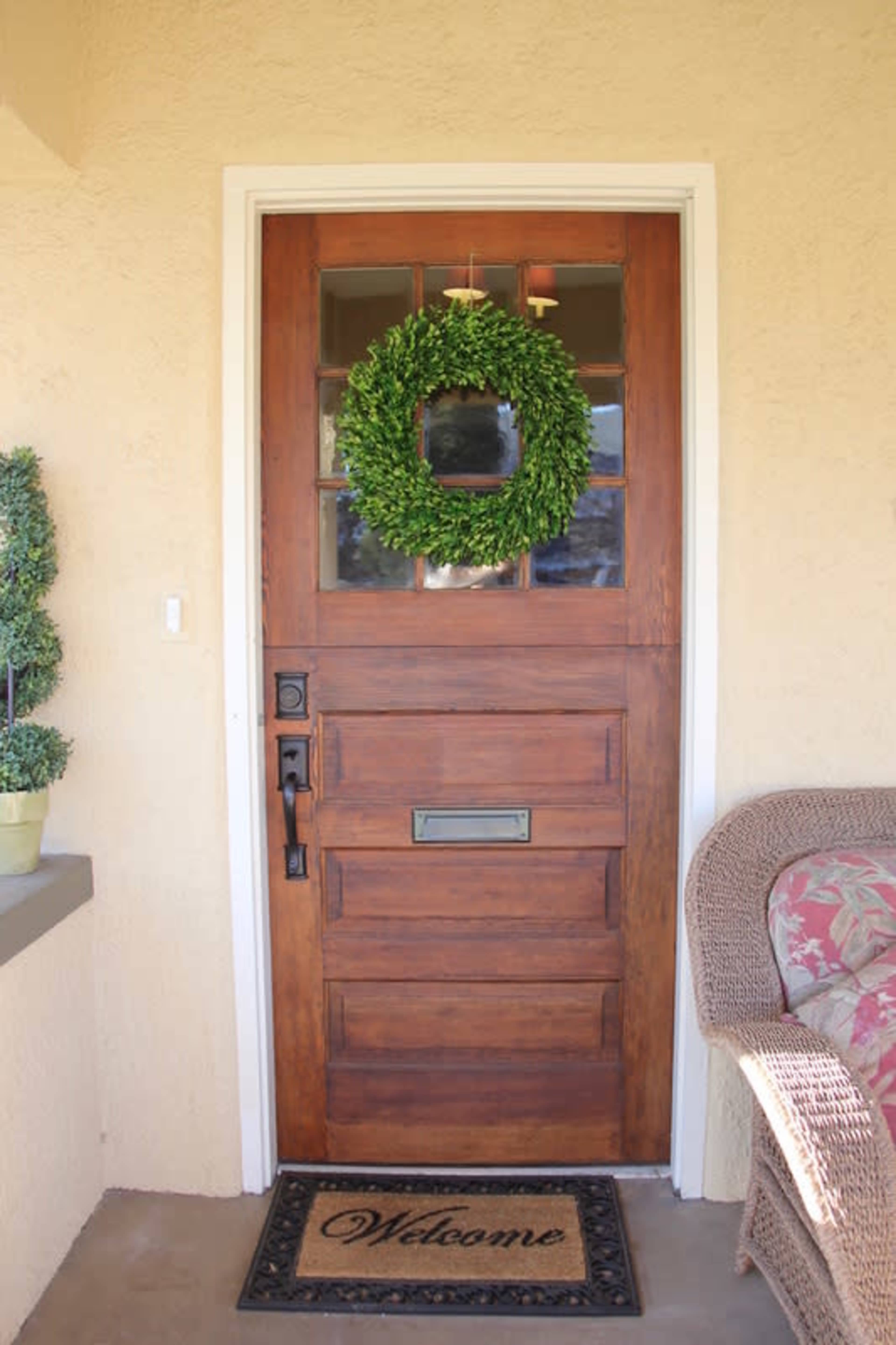 A wooden front door with a green wreath, a welcome mat, and a wicker bench beside it.