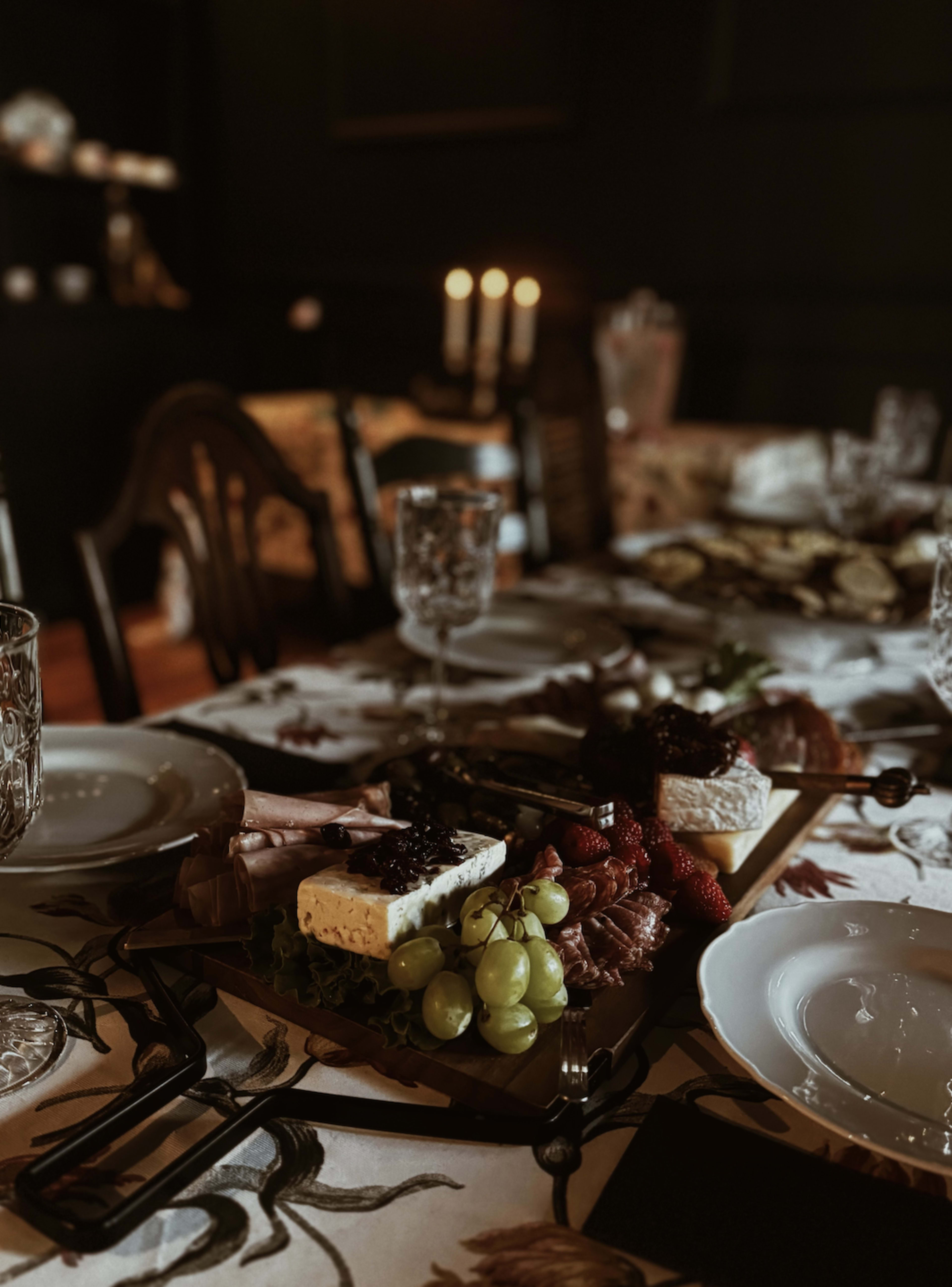 A wooden platter with various cheeses, meats, and fruits is arranged on a dining table set with crystal glassware and plates, illuminated by candlelight in a dimly lit room.