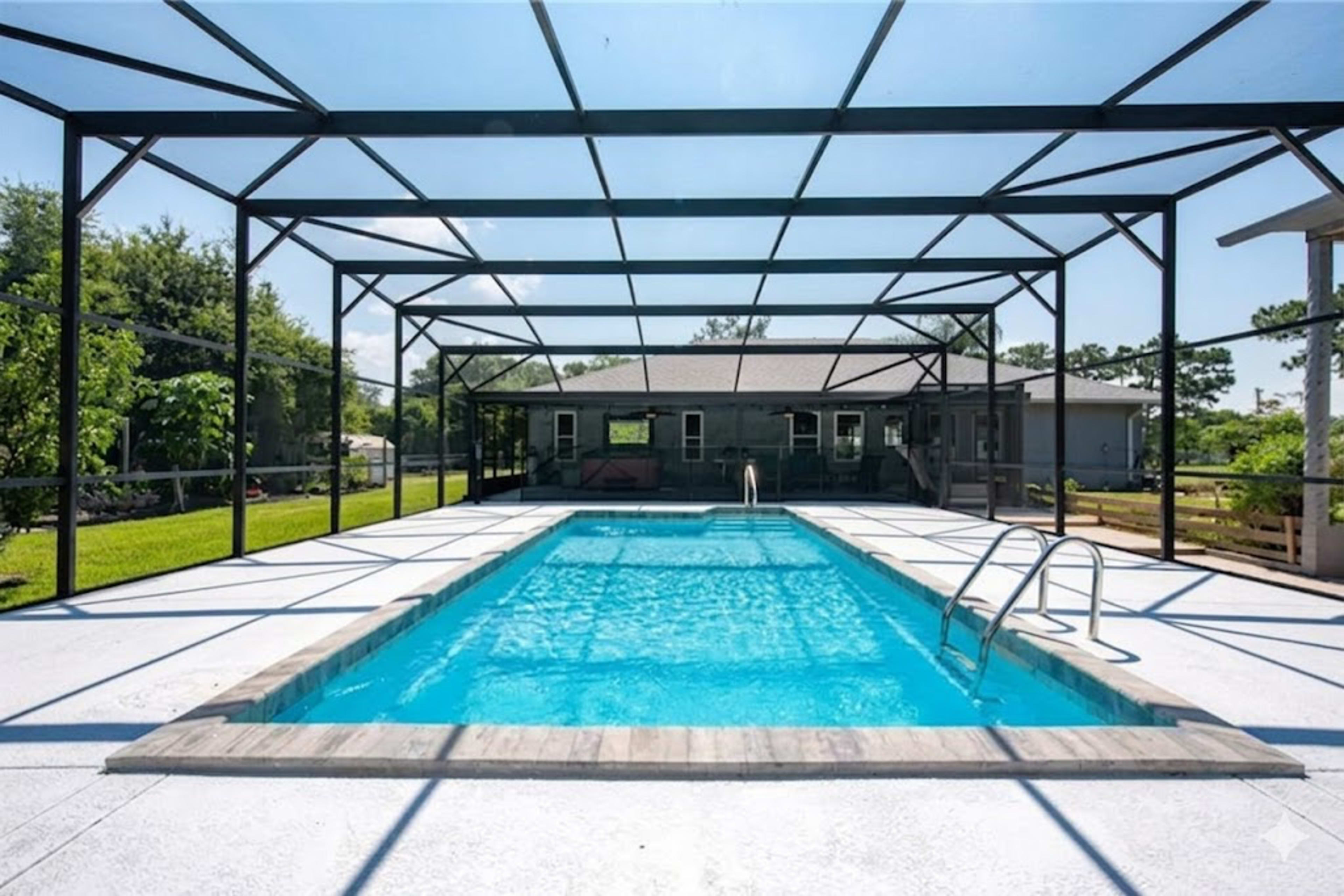 A rectangular swimming pool is enclosed by a black-frame screen, with a clear blue sky in the background and a house visible behind the pool.