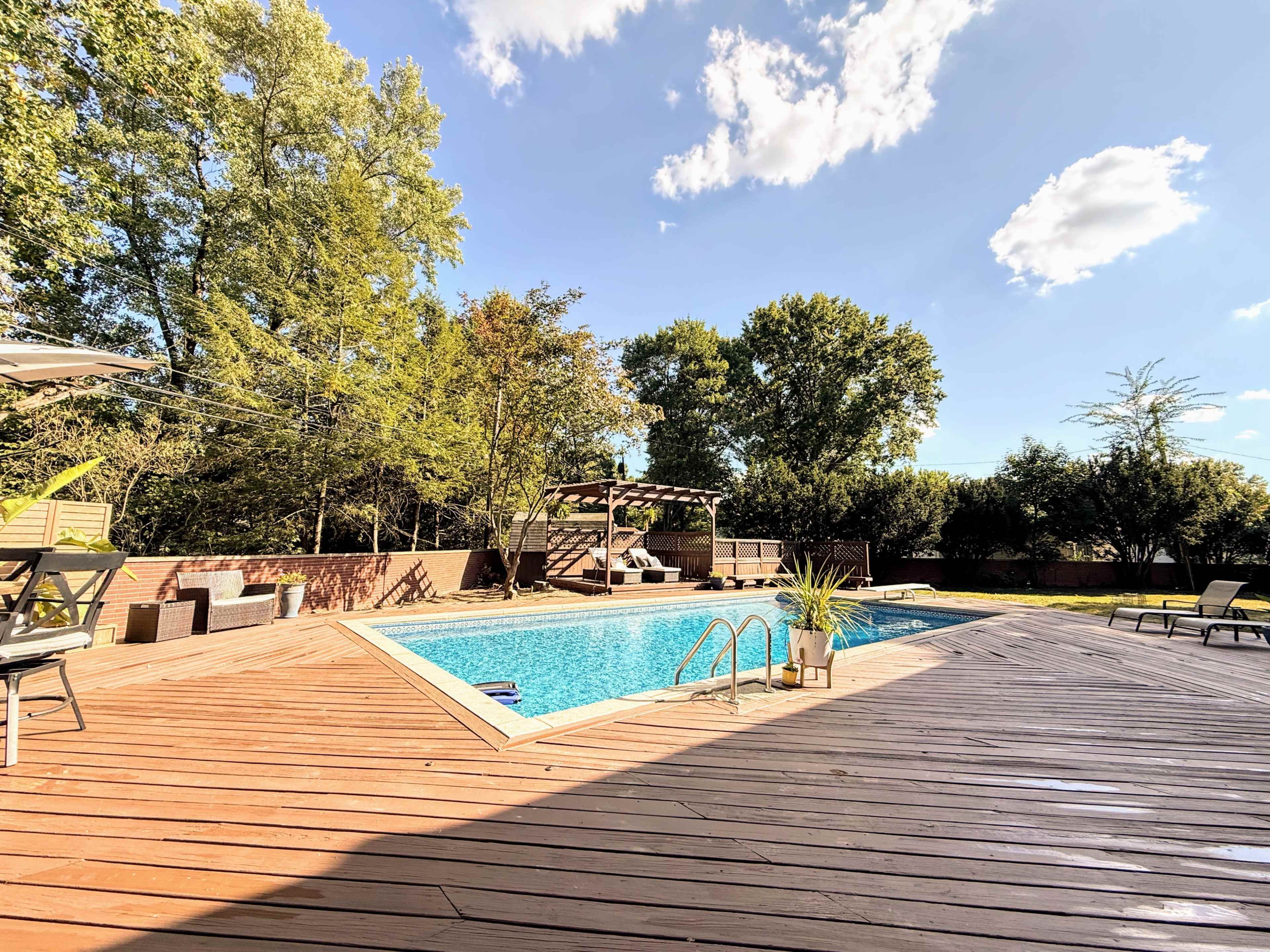The image shows a wooden deck surrounding a swimming pool with lounge chairs and a pergola in a landscaped backyard.