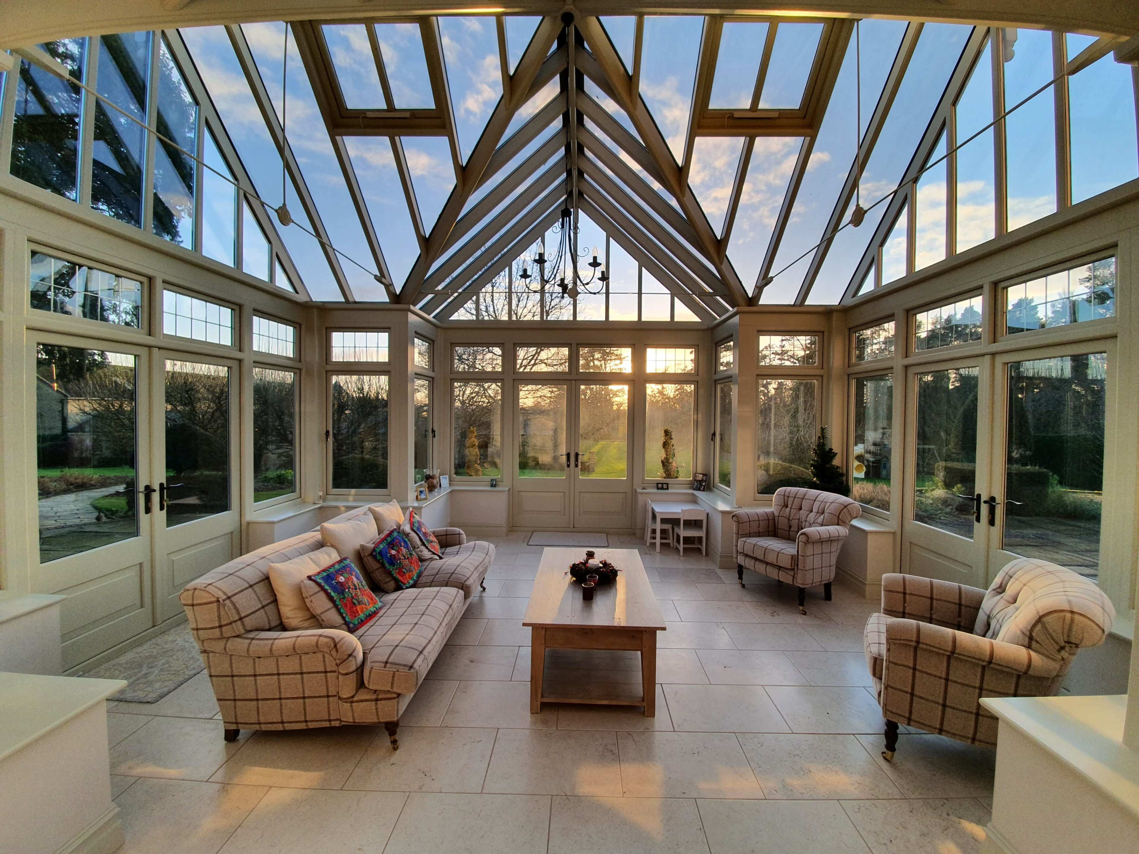 A sunlit conservatory with large glass windows, two sofas, and a coffee table set against a backdrop of trees and a sunset.