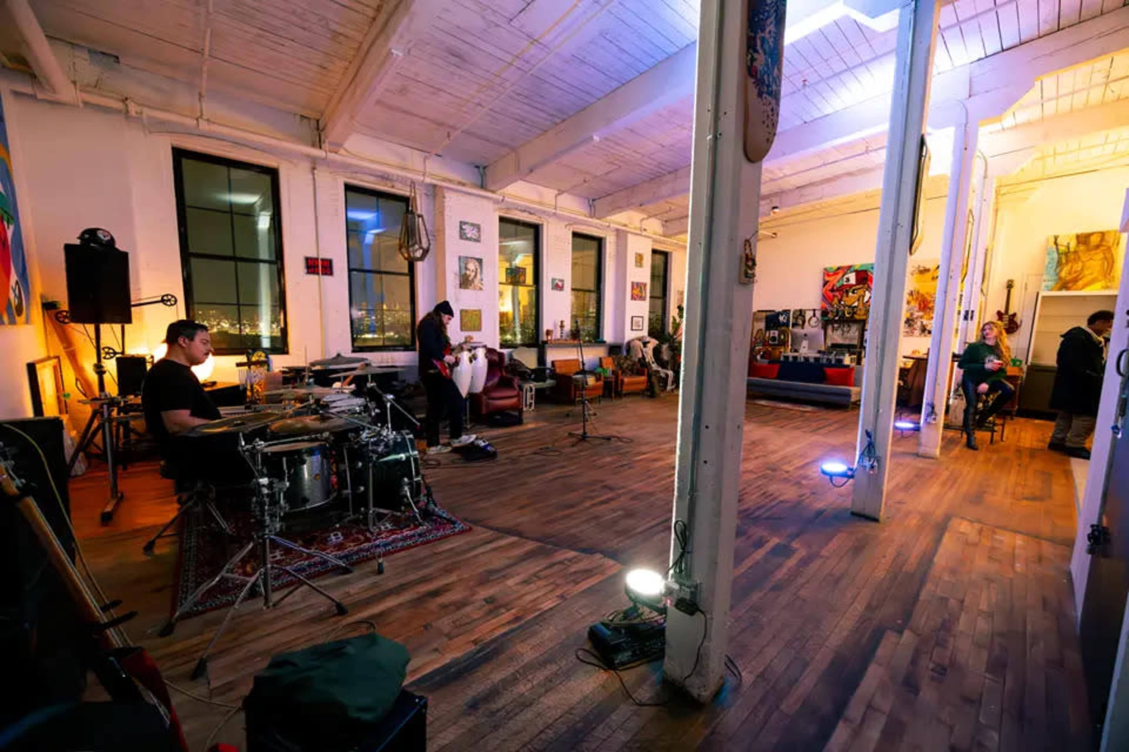 A spacious, well-lit room with wooden flooring features musicians rehearsing, including a drummer, while artwork decorates the walls.