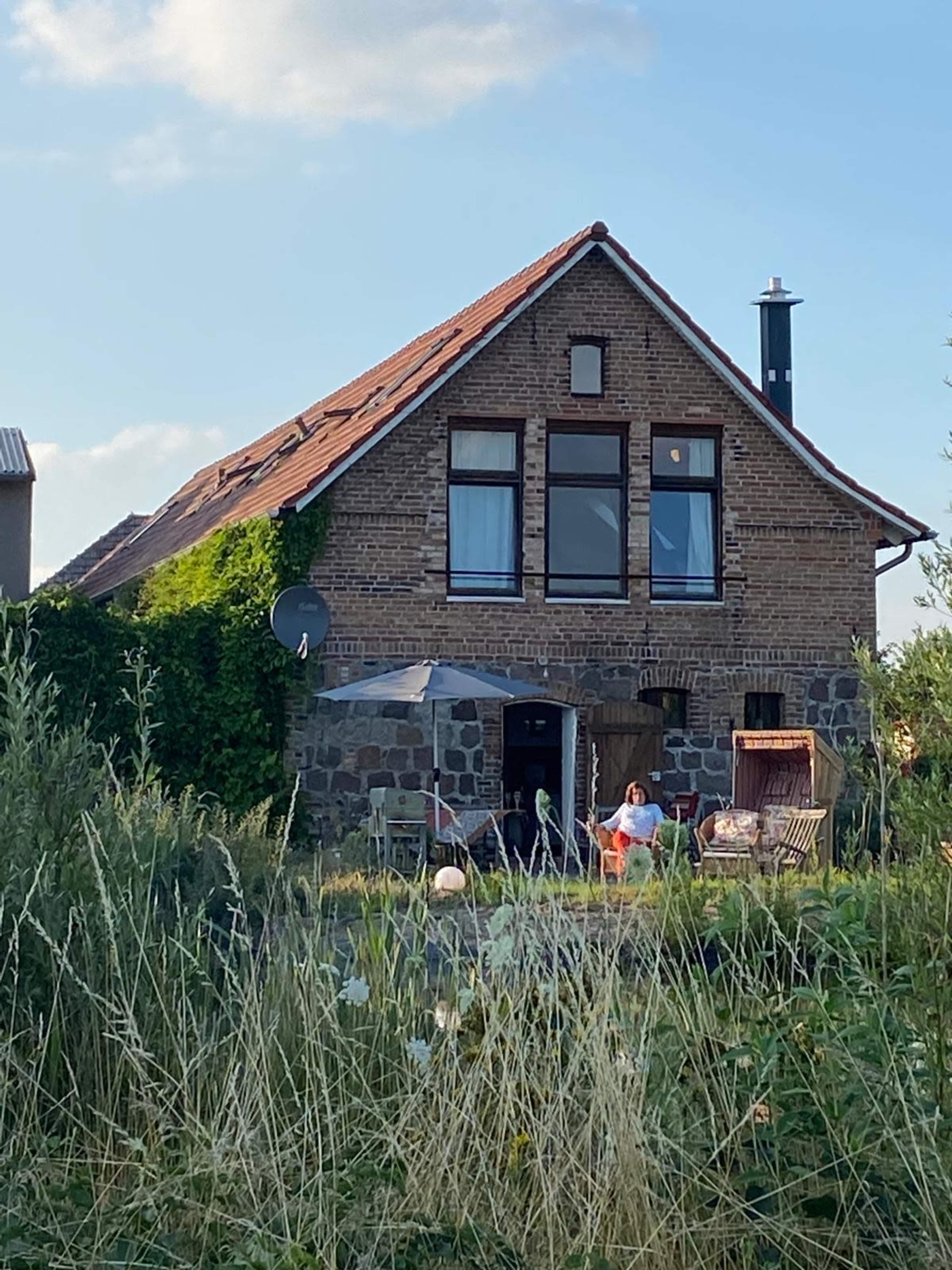 A two-story house made of stone and brick is surrounded by tall grass and plants, with outdoor seating visible in front.