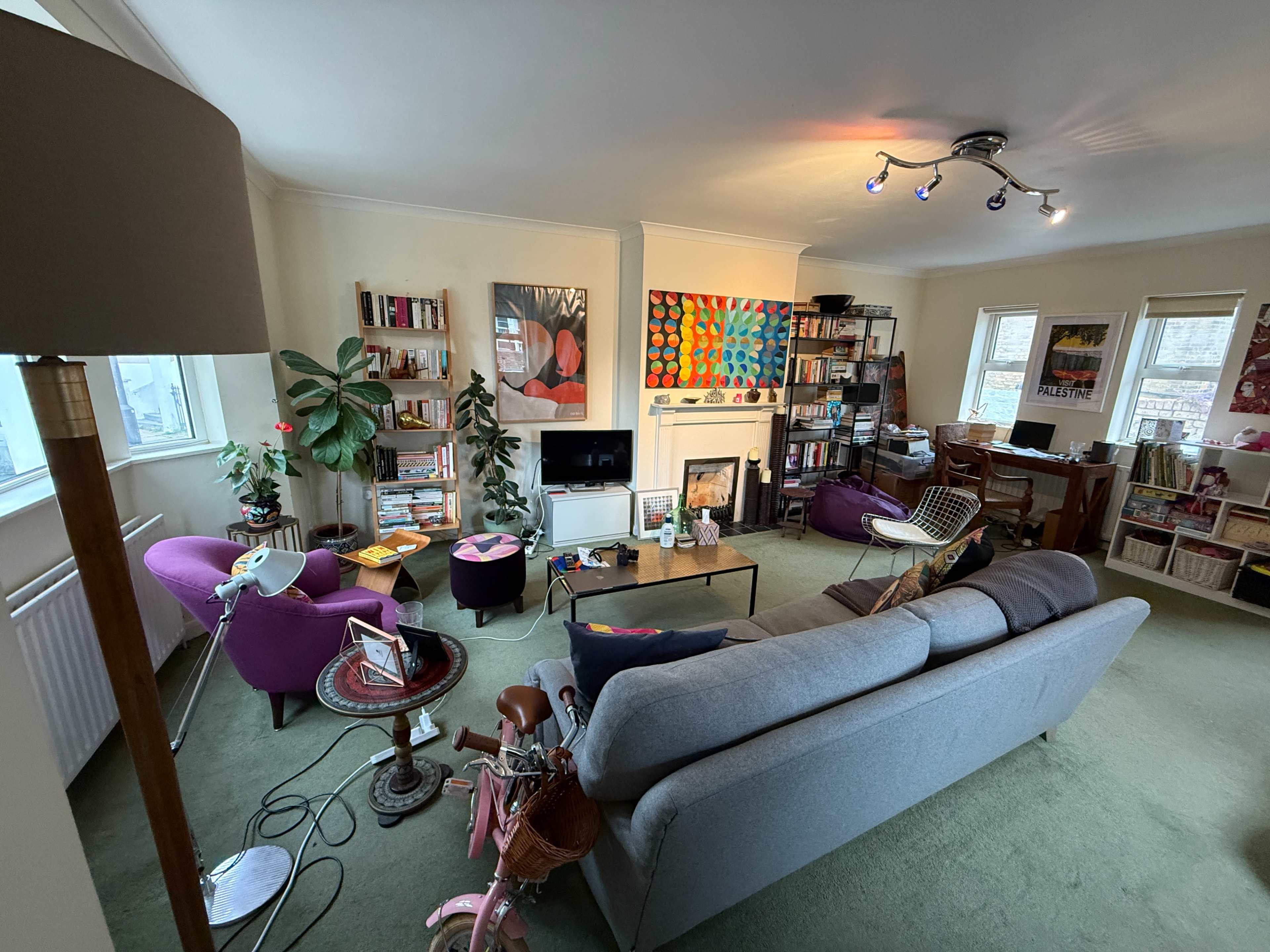 The image shows a cozy living room with a gray sofa, purple armchairs, a coffee table, bookshelves filled with books, and various decorations on the walls.