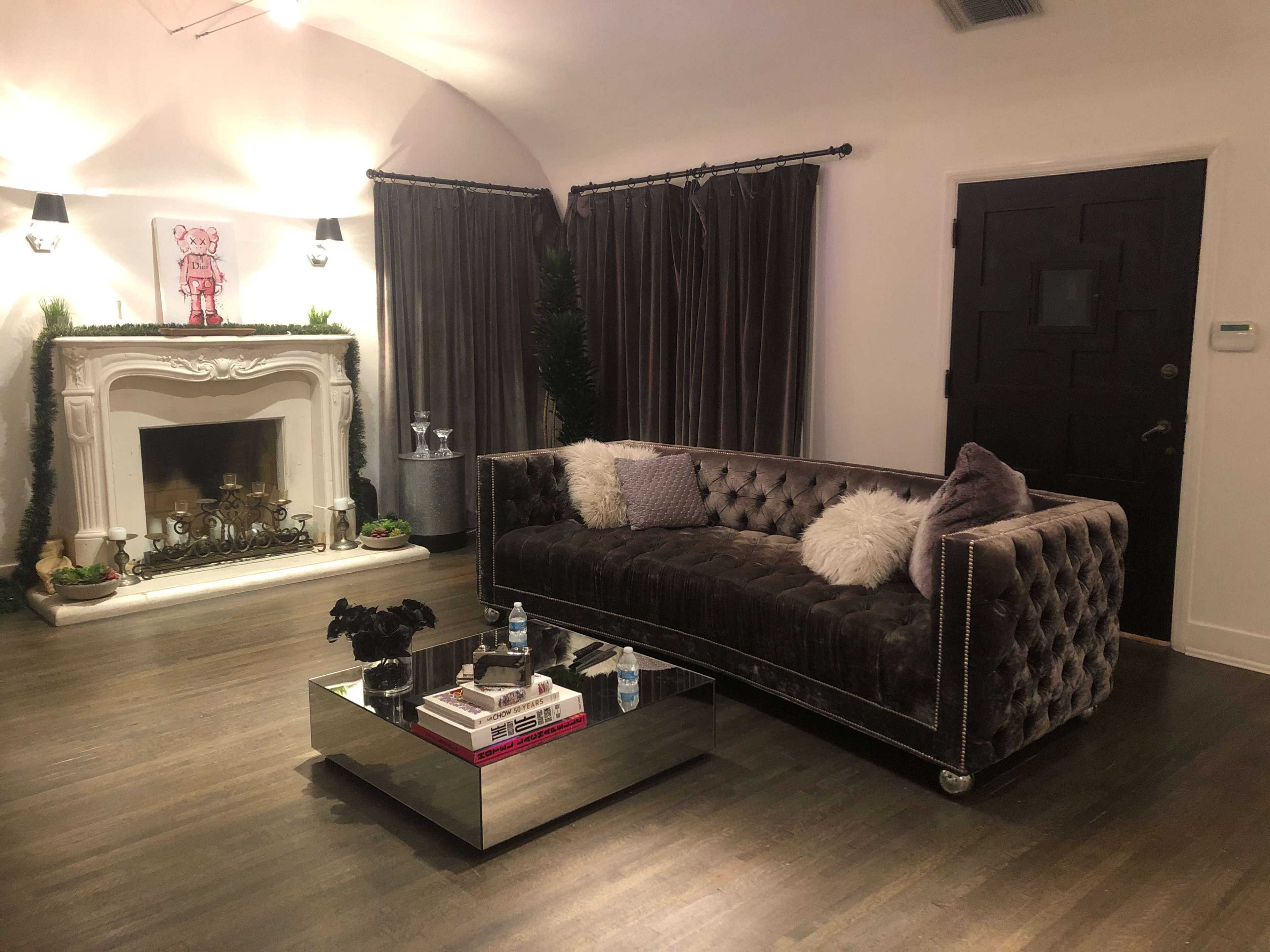 The room features a velvet sofa, a coffee table, a decorative fireplace, and dark curtains, creating a cozy living space.