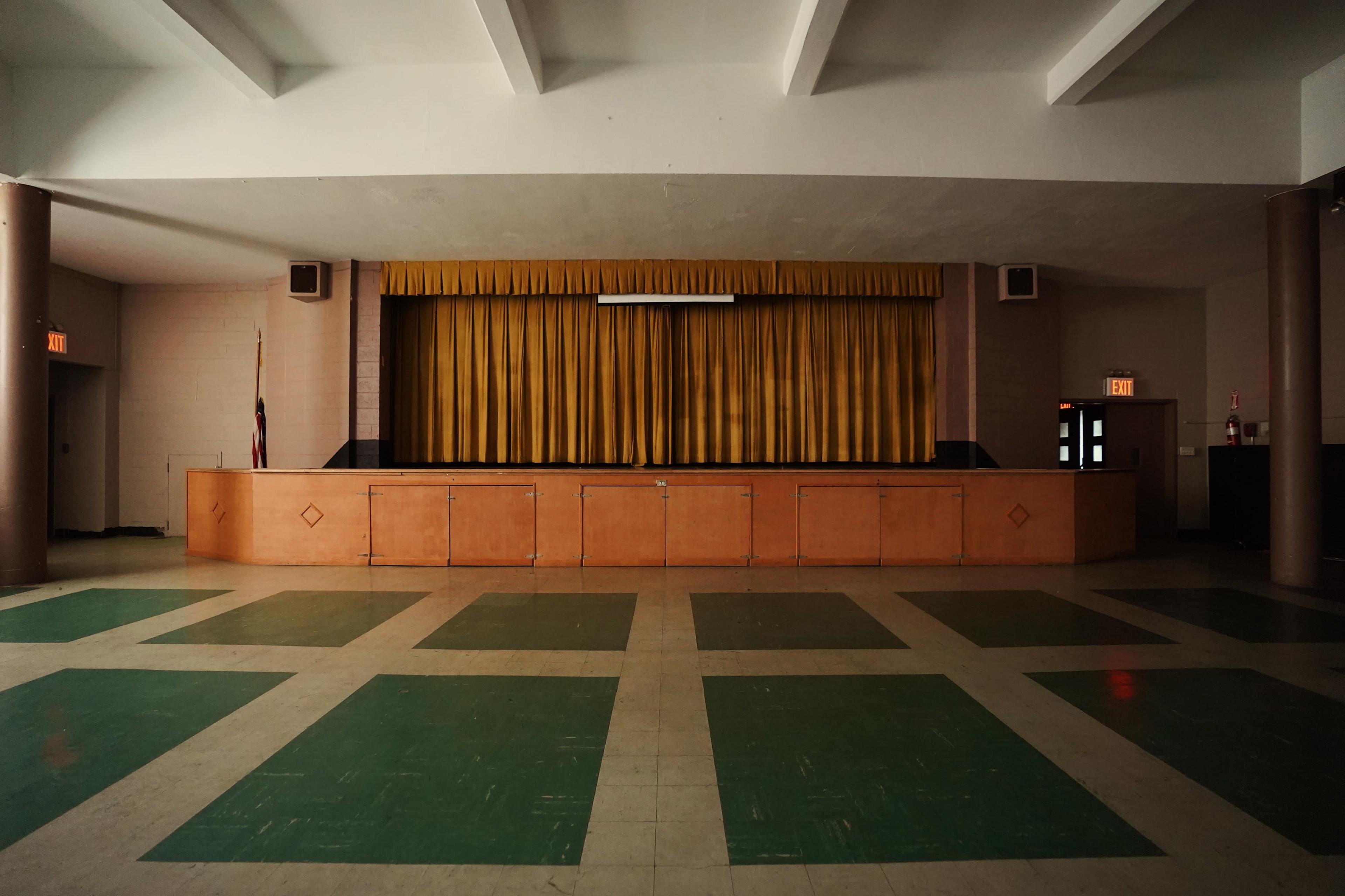 An empty stage with yellow curtains, set in a large room with a tile floor featuring a green and beige checkerboard pattern.