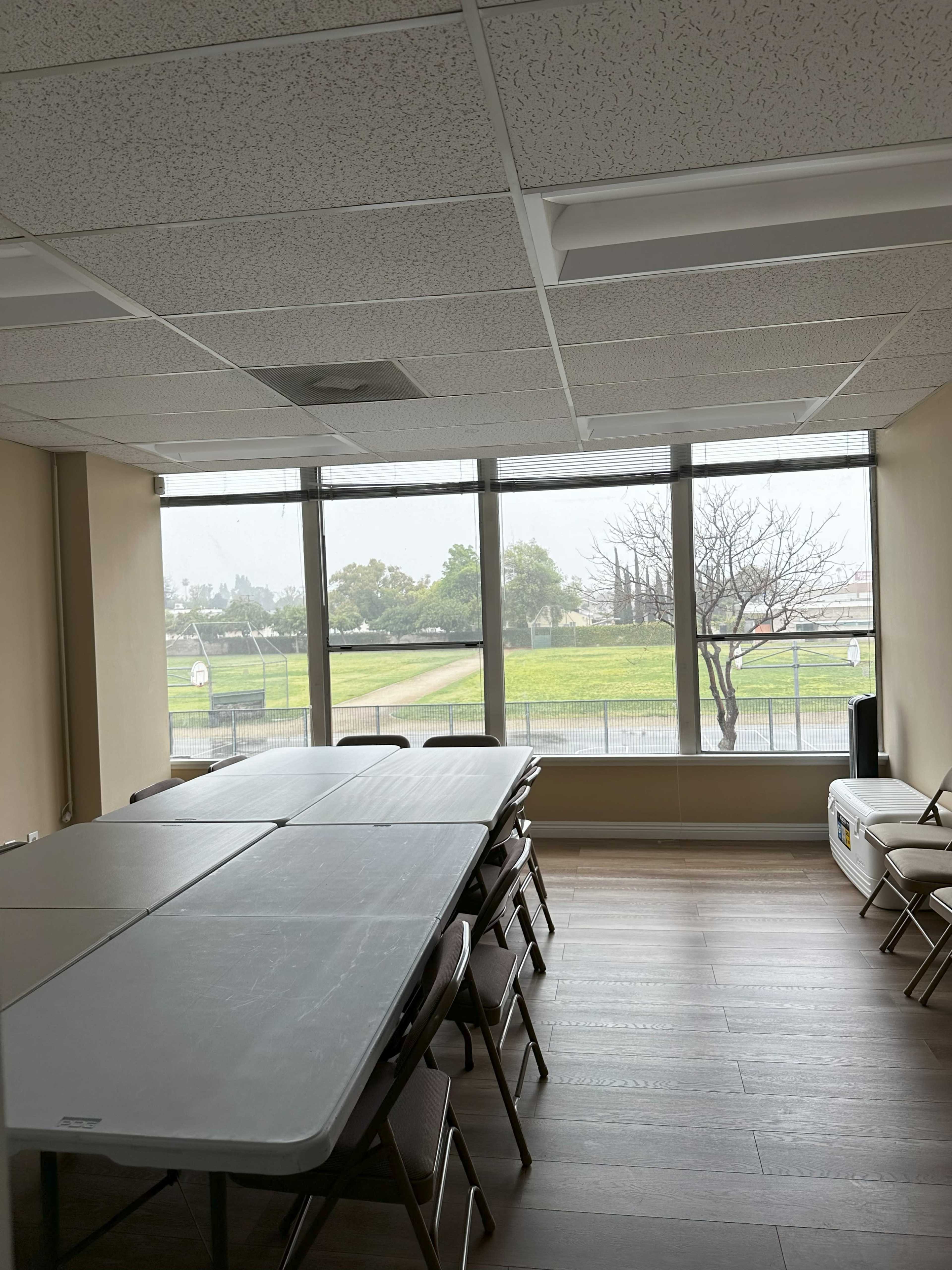 A large room with a rectangular table and chairs is illuminated by natural light coming through wide windows, overlooking a grassy area and a pathway.