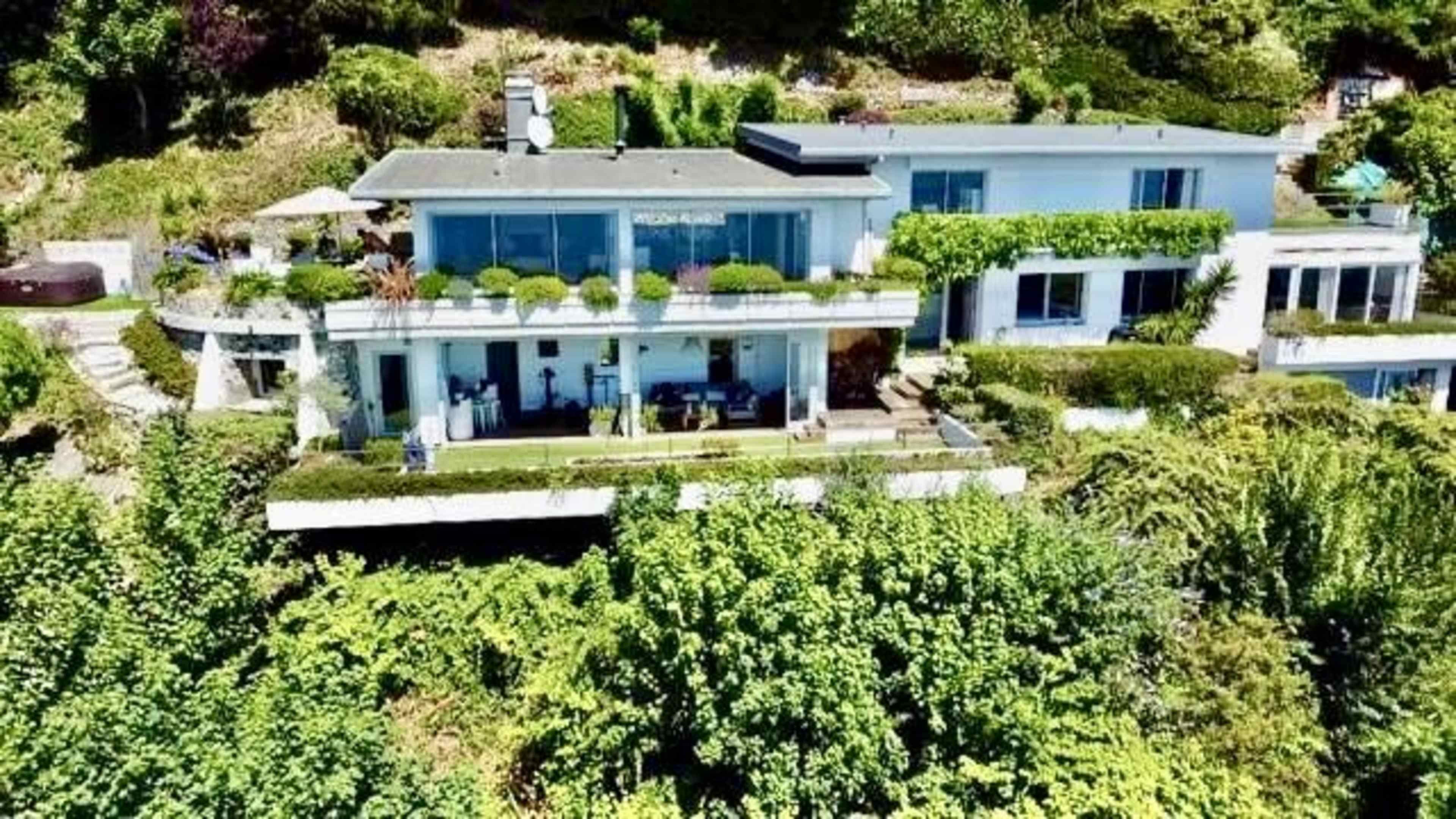 A modern hillside house with large windows is surrounded by greenery and features multiple levels of terraces.