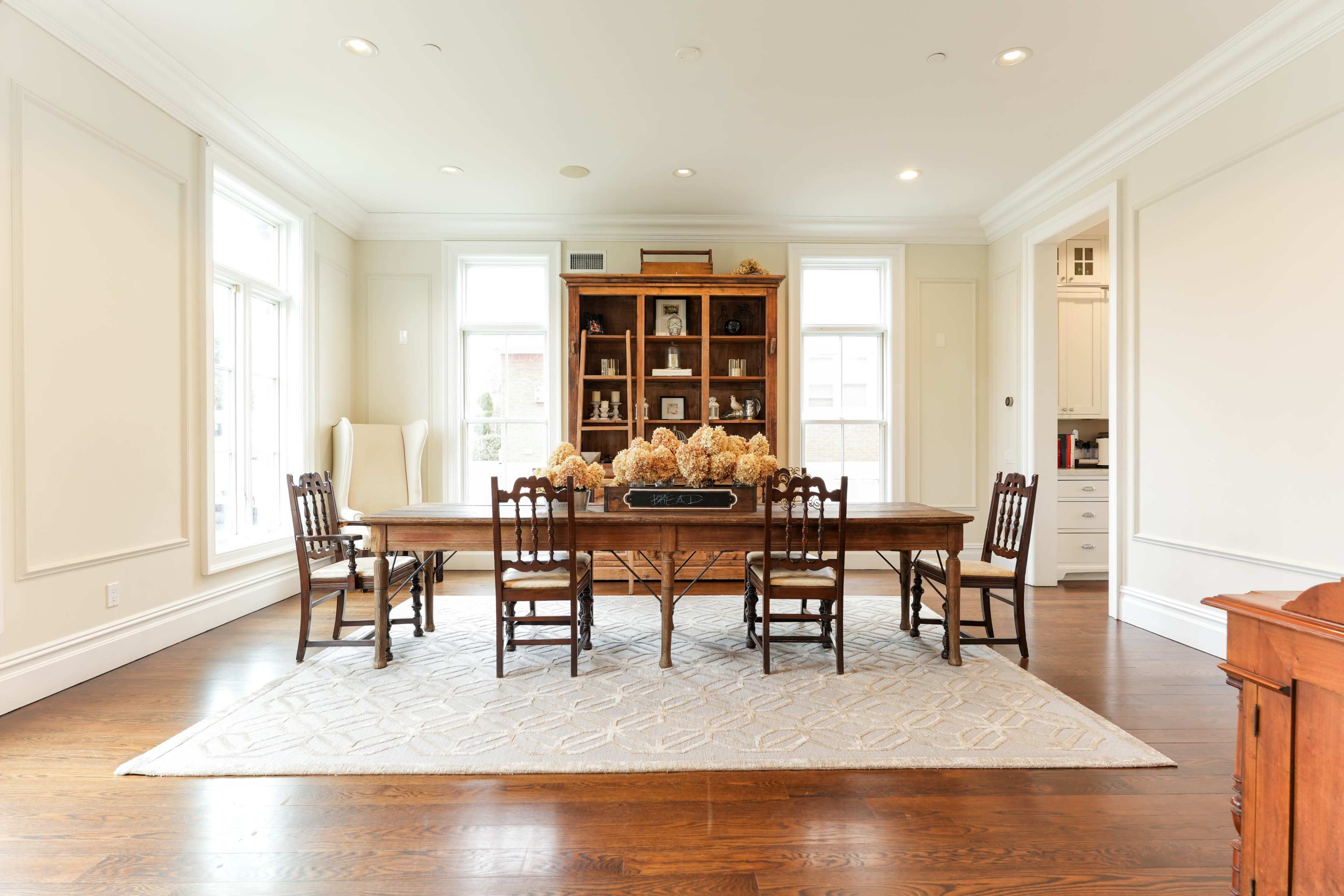 A wooden dining table surrounded by chairs is set in a well-lit room with large windows and a display cabinet in the background.