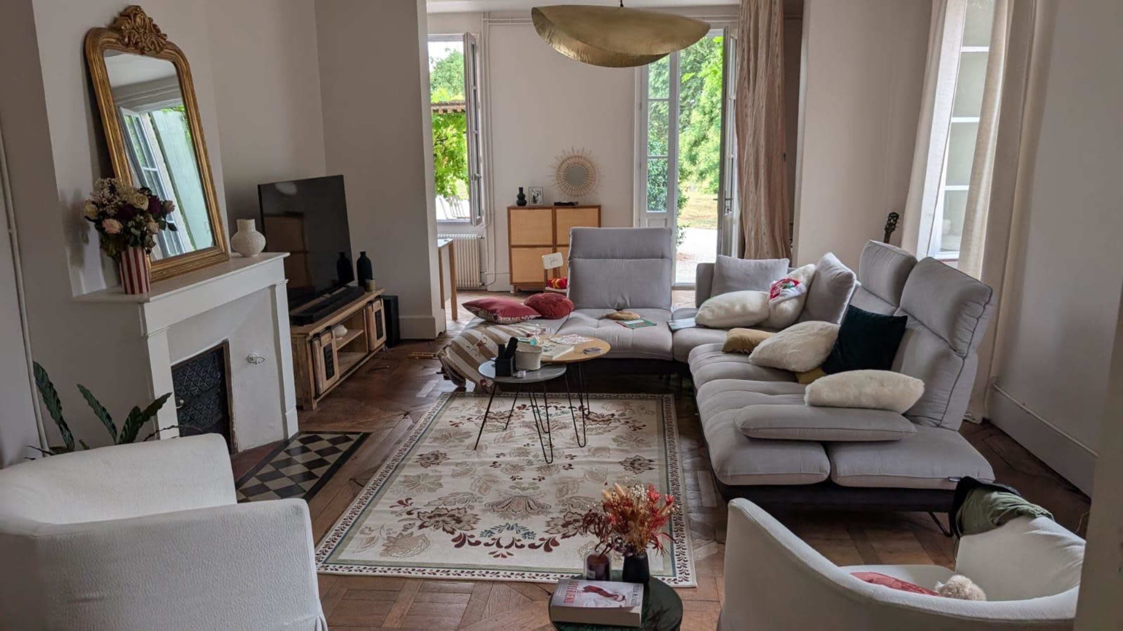 A living room features a sectional sofa, two armchairs, a coffee table, and a large area rug, illuminated by natural light from the windows.