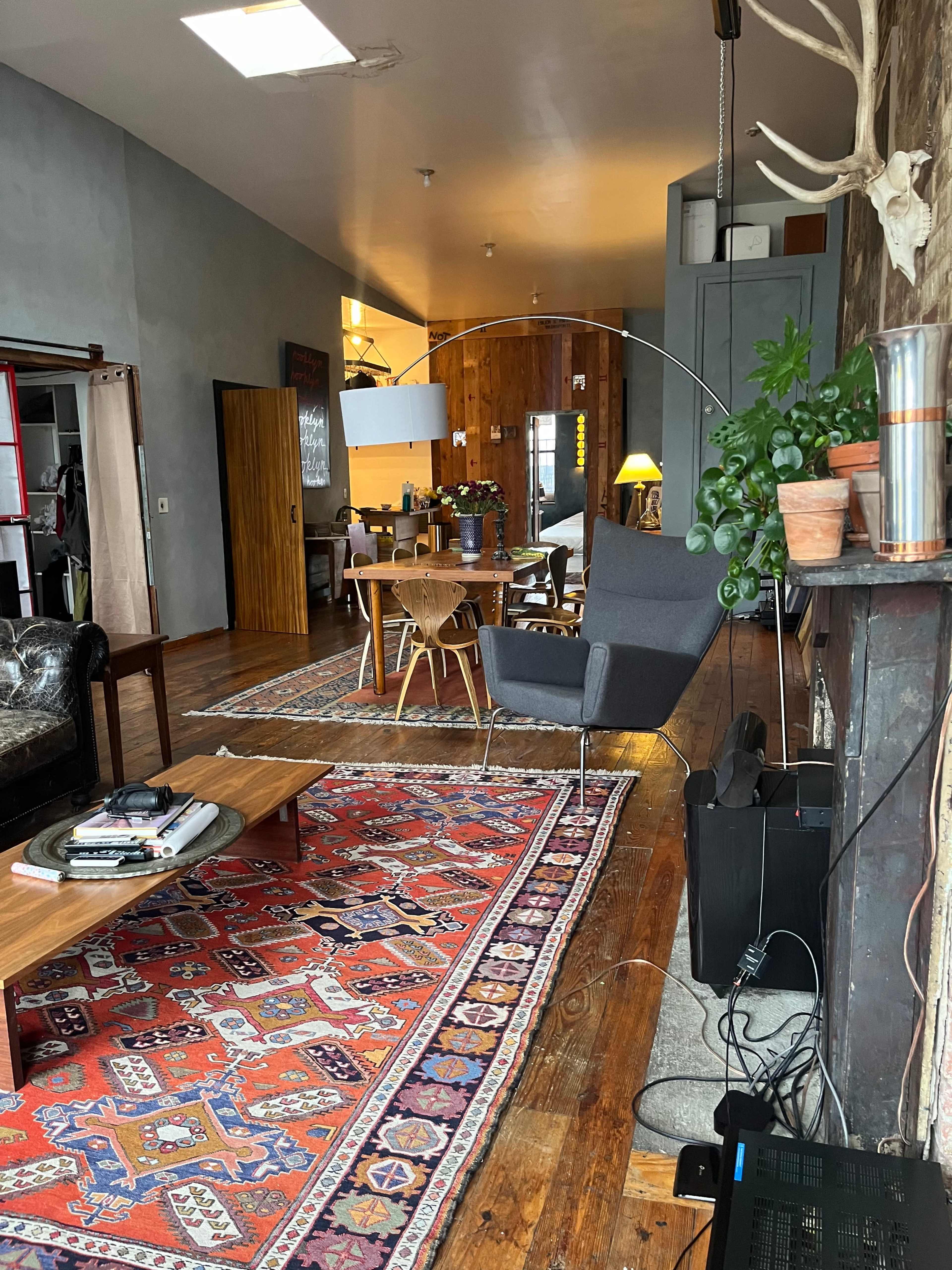 A spacious, modern living area with wooden floors, a large area rug, a seating area with a gray chair, a coffee table, and a dining space visible in the background.