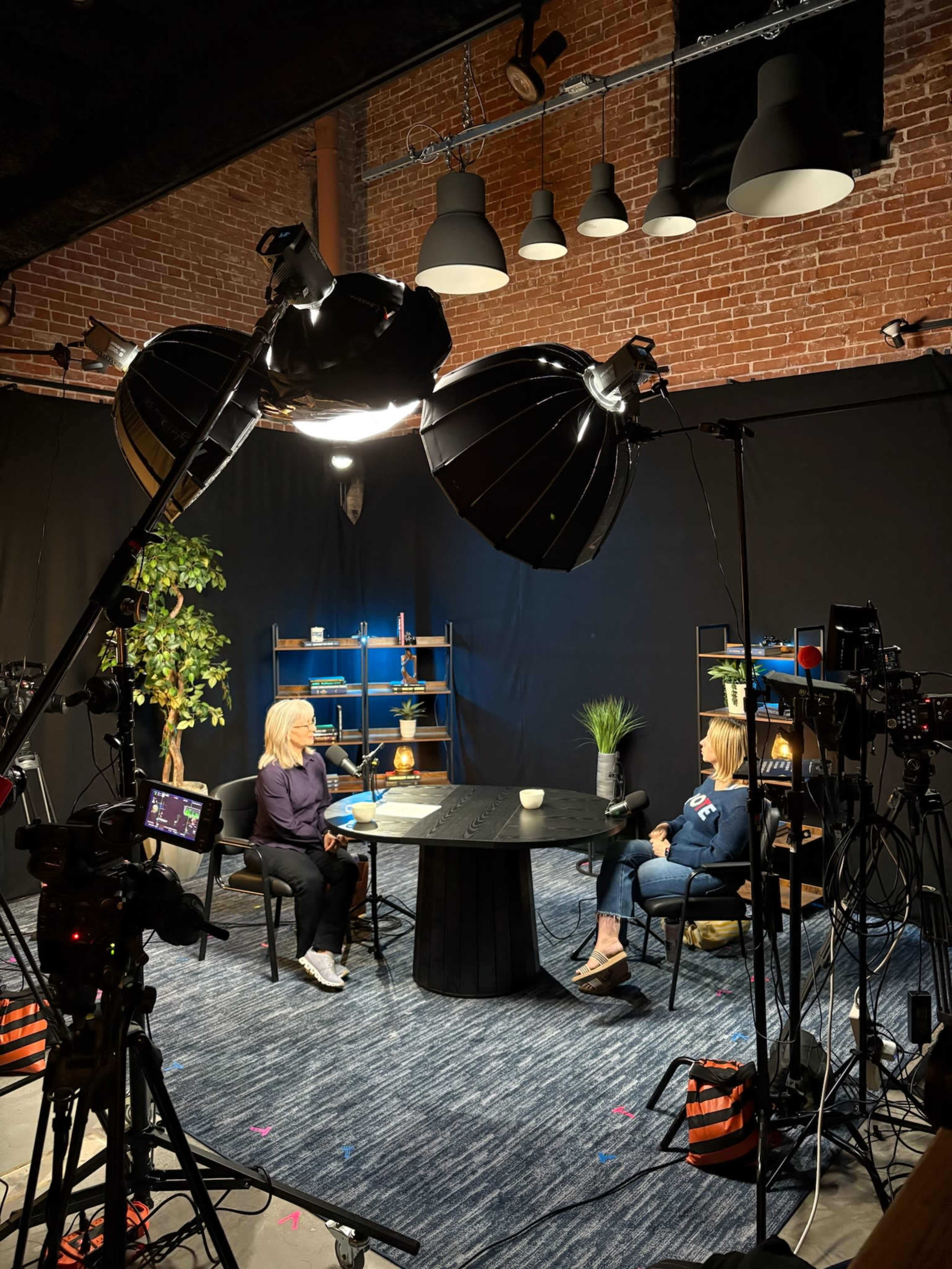 The image shows two individuals seated at a table in a studio setting, surrounded by various filming equipment and decorative elements including a potted plant and shelves.