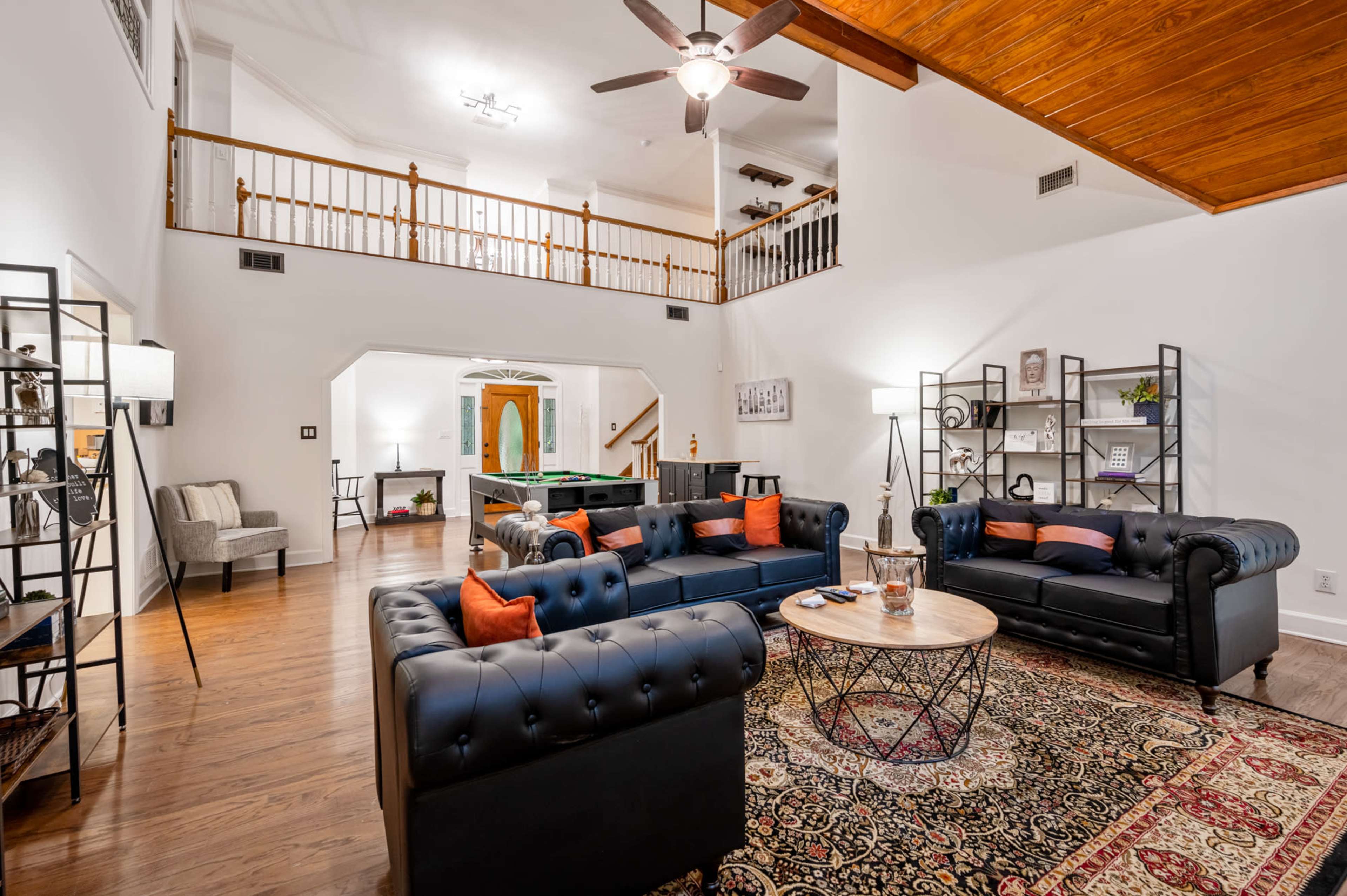 A spacious living room features two black leather couches, a round coffee table, and a decorative rug, with a view of a second-level balcony and an open area leading to a game room in the background.